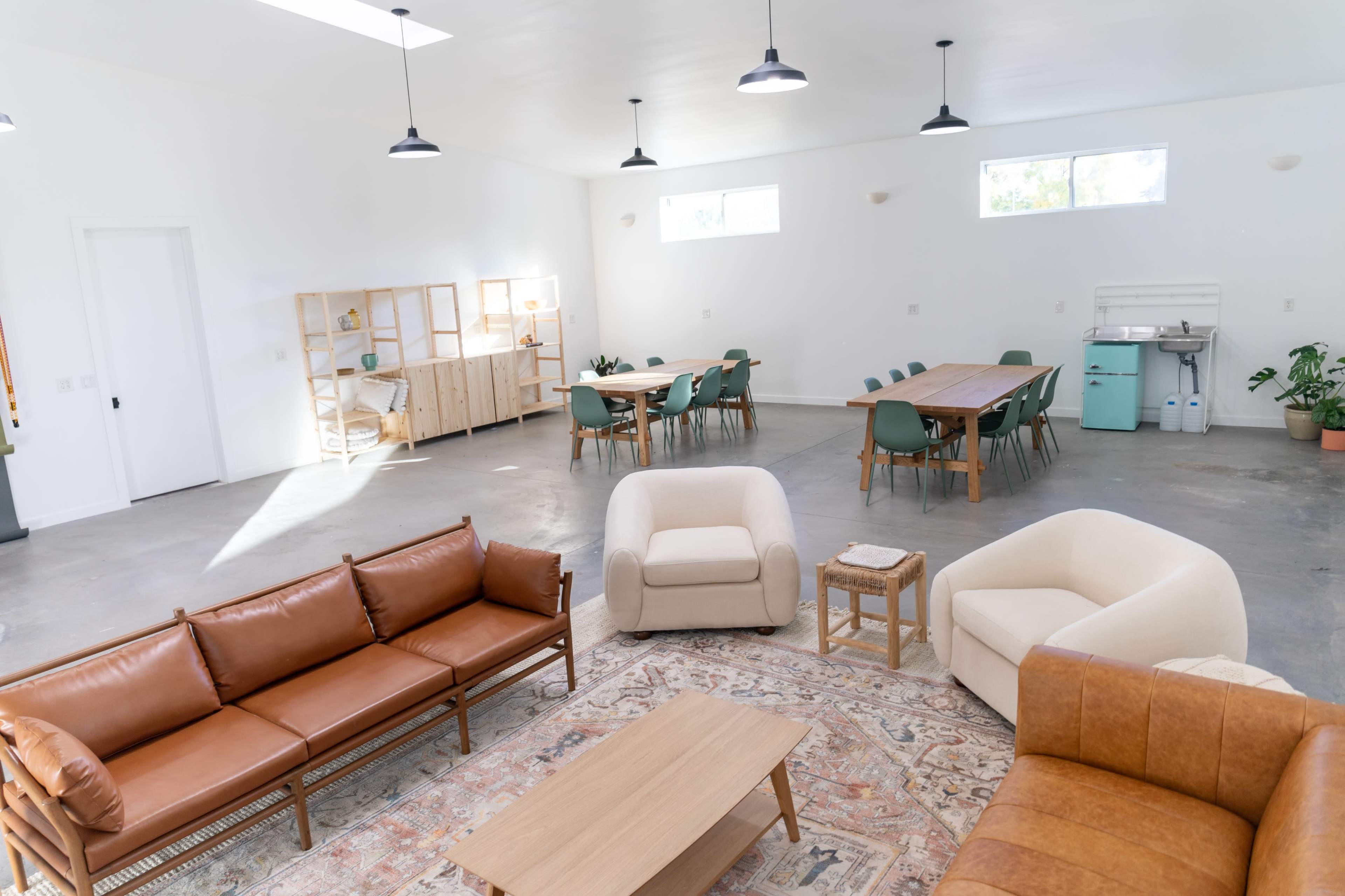 A spacious, modern room with a seating area featuring brown and cream sofas, a wooden coffee table, and a dining area with green chairs and a wooden table under pendant lighting.