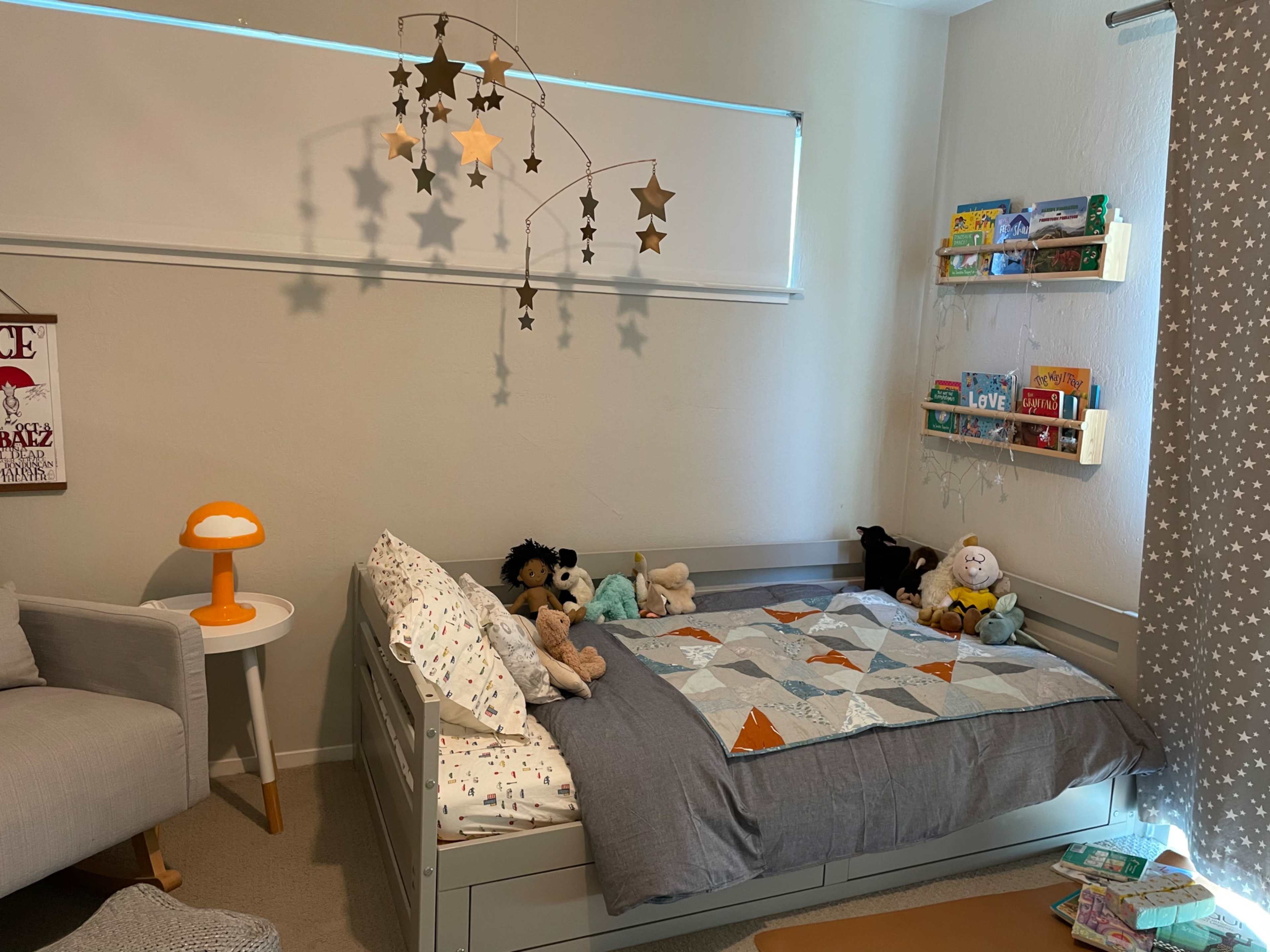 A cozy children's bedroom features a bed with a colorful quilt, a lamp beside it, a mobile of stars hanging above, and shelves filled with books and toys.
