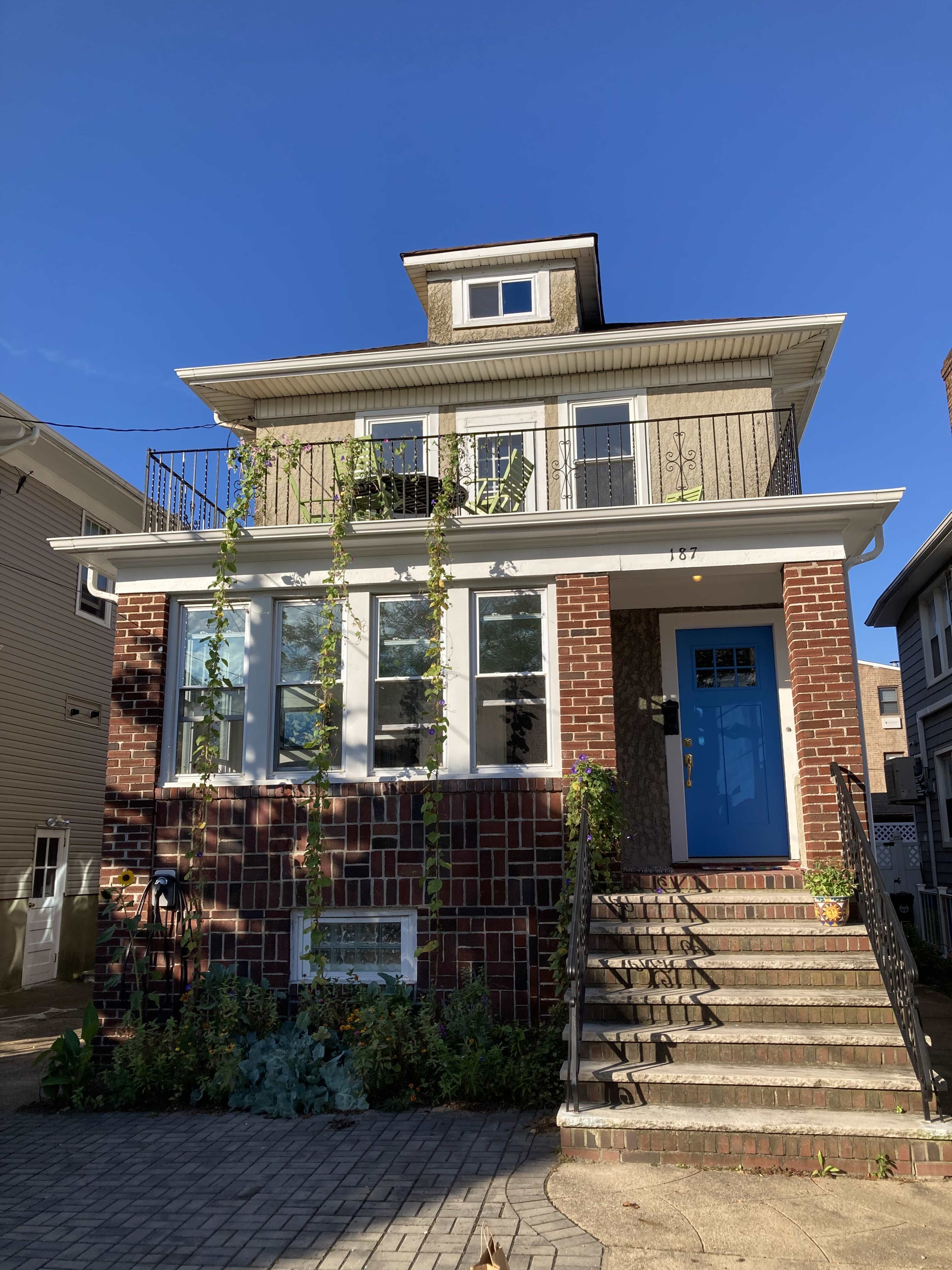 A three-story brick house with a blue door features a small balcony and a staircase leading to the entrance, surrounded by greenery.