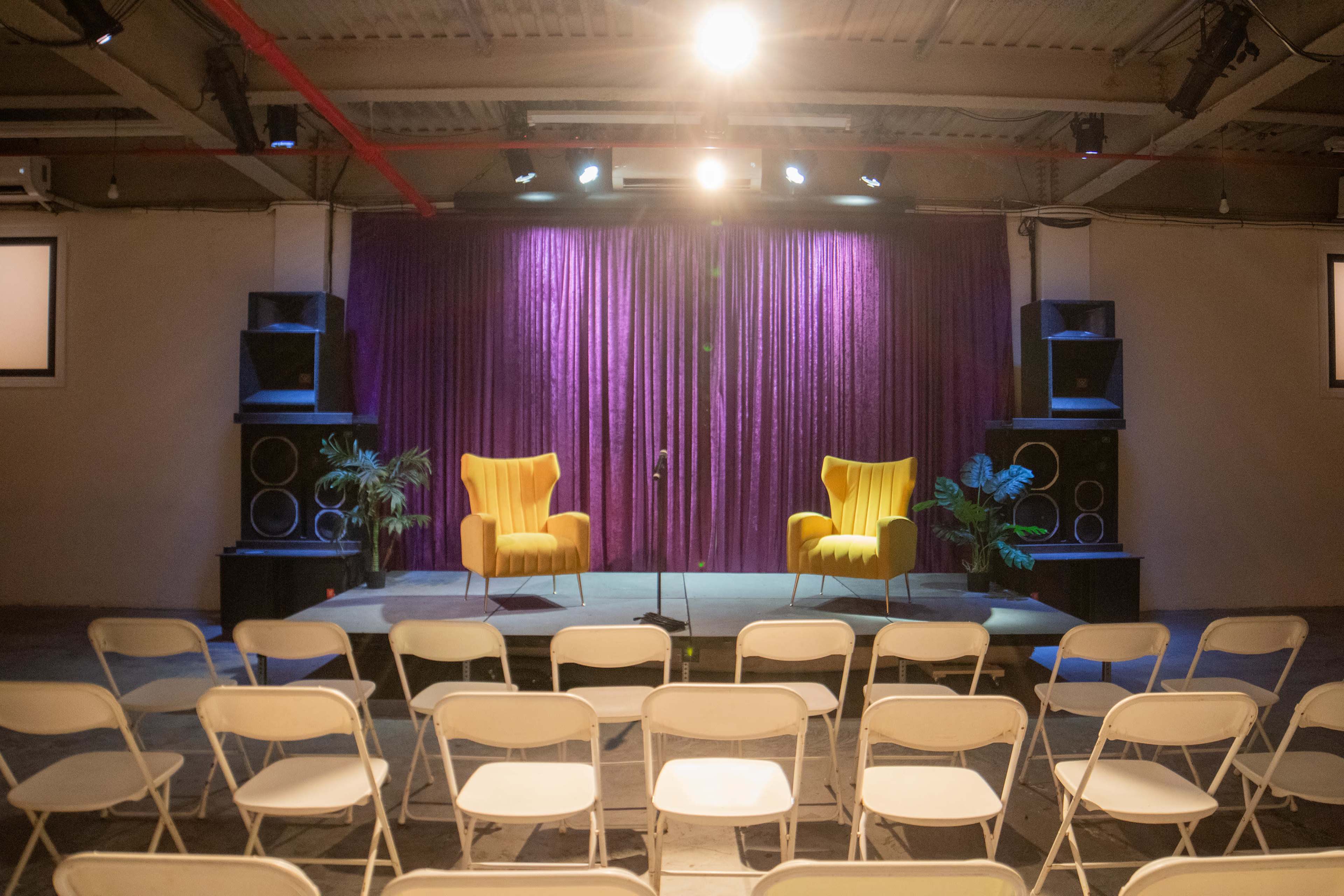 A stage with two yellow chairs set against a purple curtain, flanked by large speakers, and facing a row of white folding chairs.