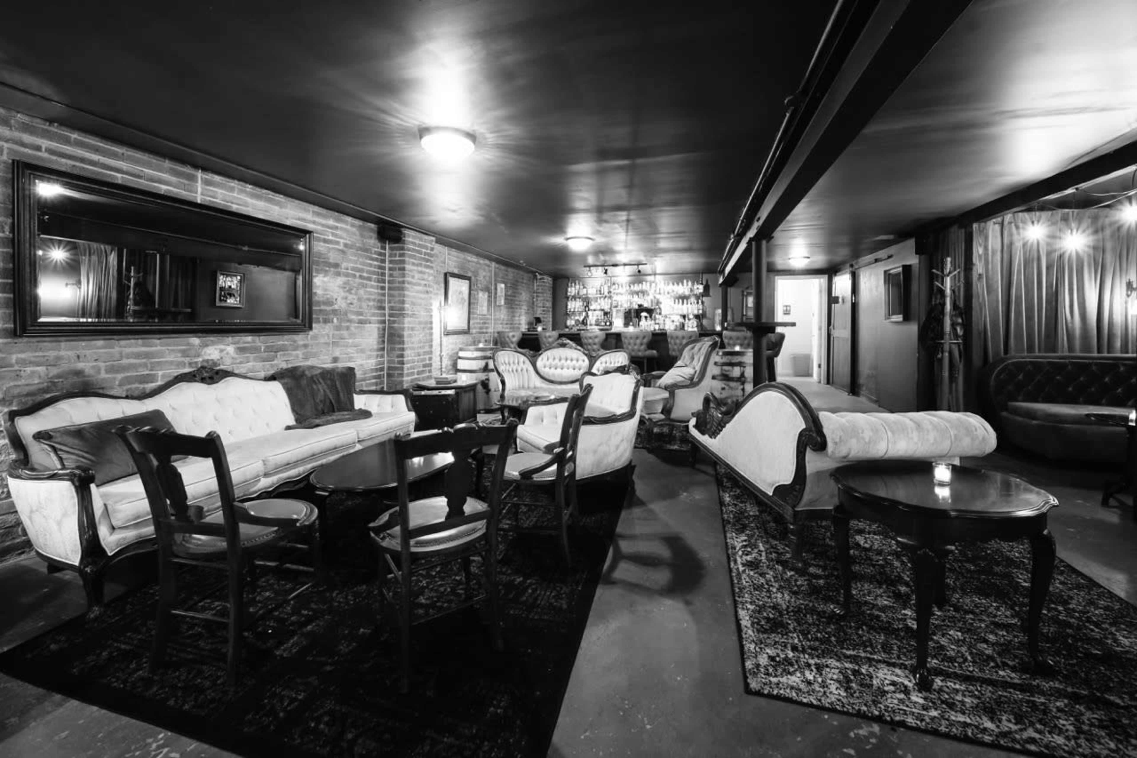 The image depicts a dimly lit lounge with vintage furniture, including sofas and chairs arranged around small tables, and a bar area visible in the background.