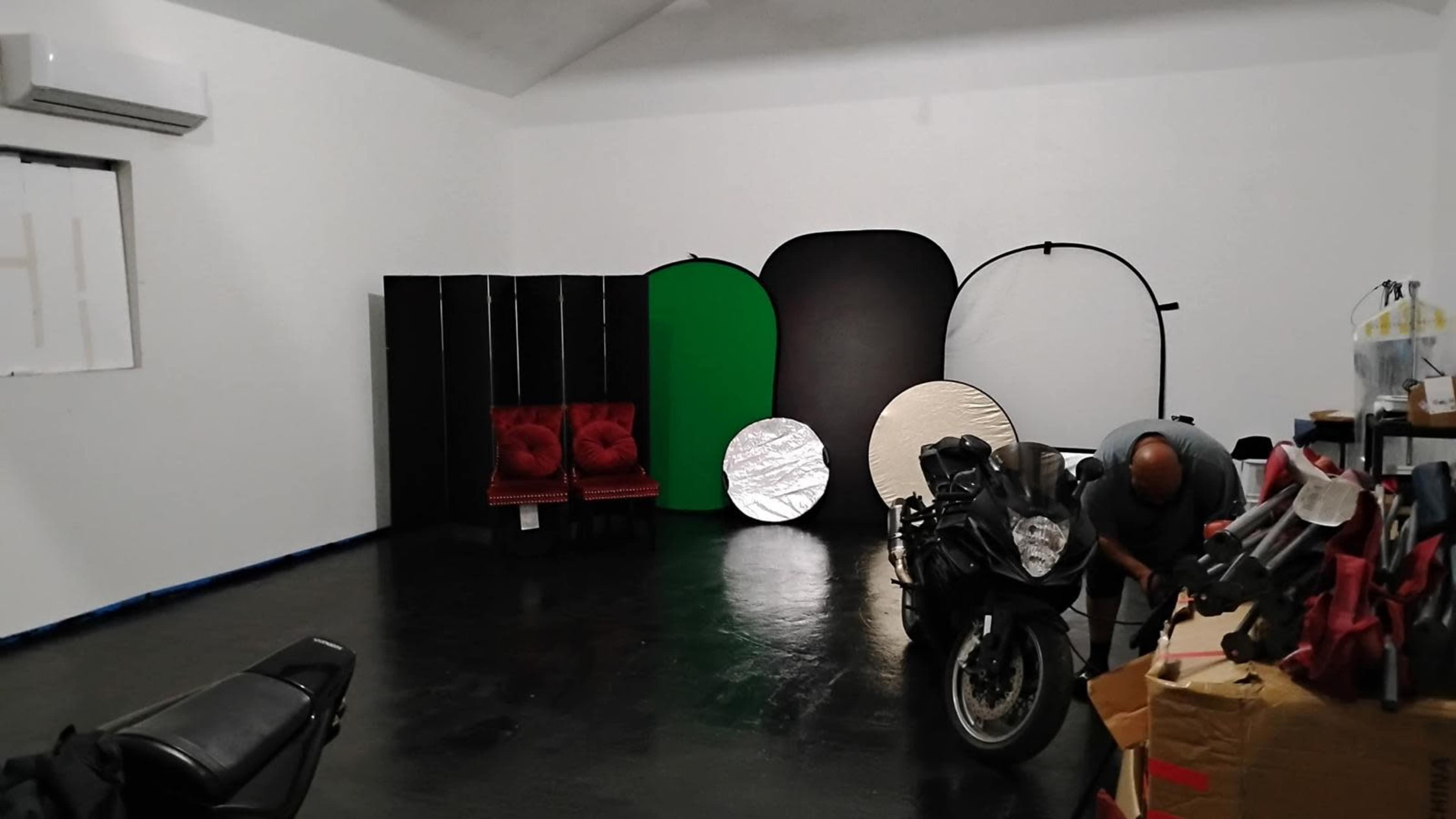 The image shows a spacious room with a black floor, two red chairs, several backdrop panels in various colors, and a motorcycle near a stack of items.