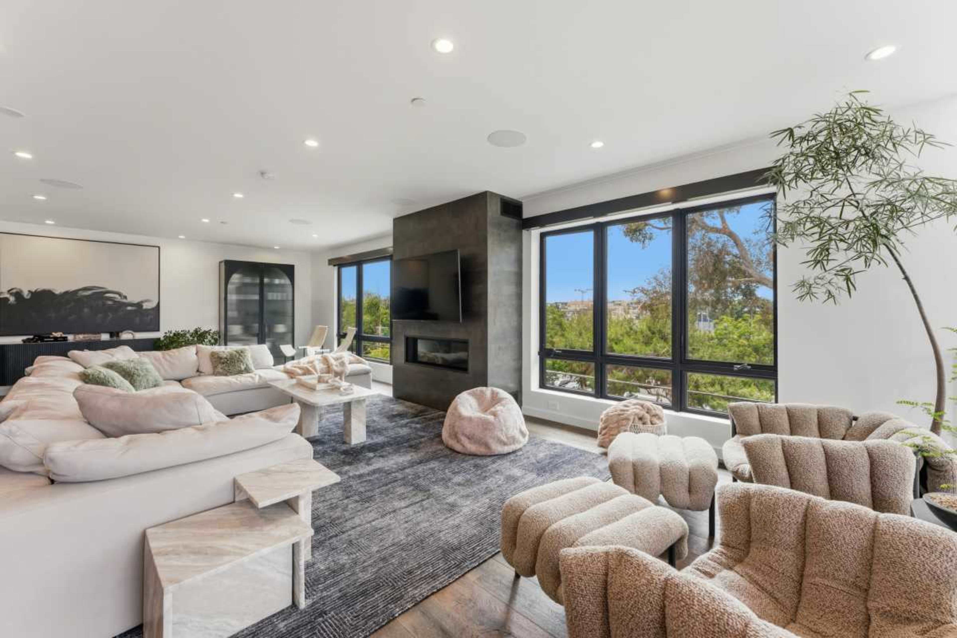 The image displays a modern living room with a large sectional sofa, armchairs, a coffee table, and large windows providing a view of greenery outside.