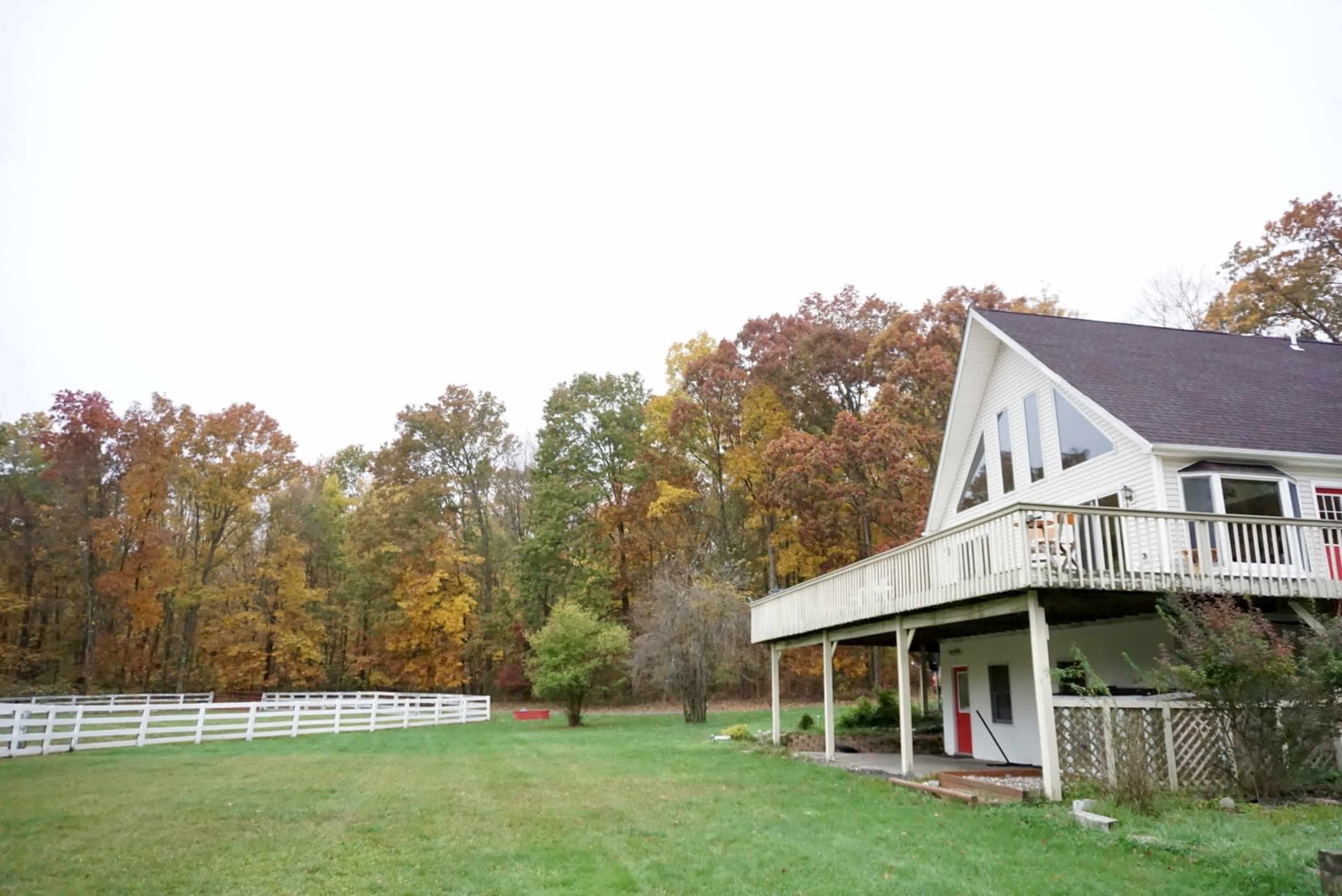 A two-story house with a deck is situated next to a grassy area and a white fence, bordered by trees in autumn colors.