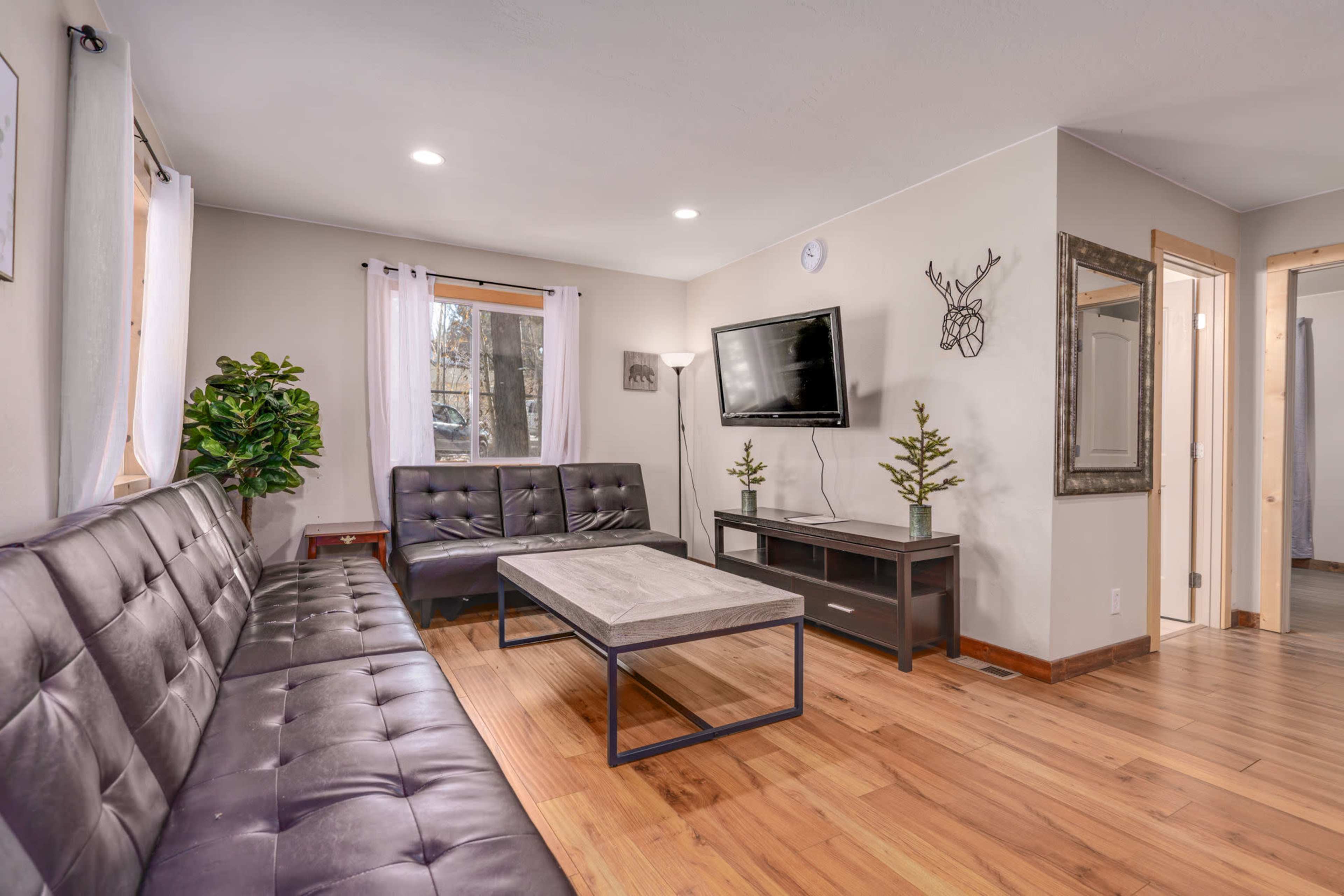 A modern living room features a dark leather sofa, a coffee table, and a TV mounted on the wall, with natural light coming through the windows.