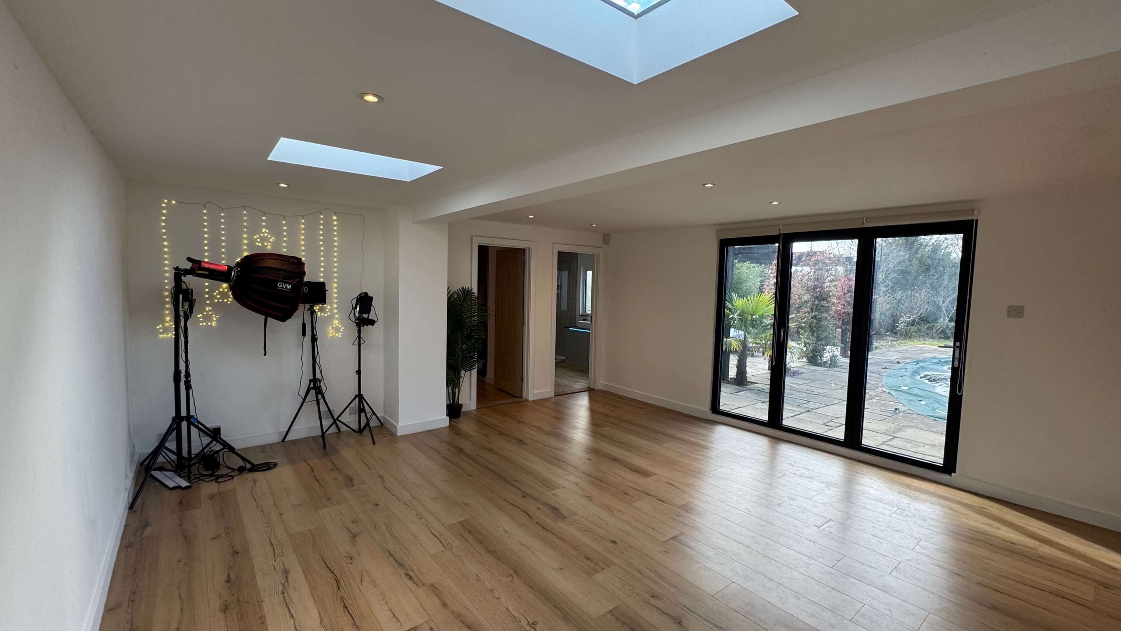 The image shows a spacious, well-lit room with wooden flooring, featuring a set of lighting equipment and large glass doors leading to a garden area.