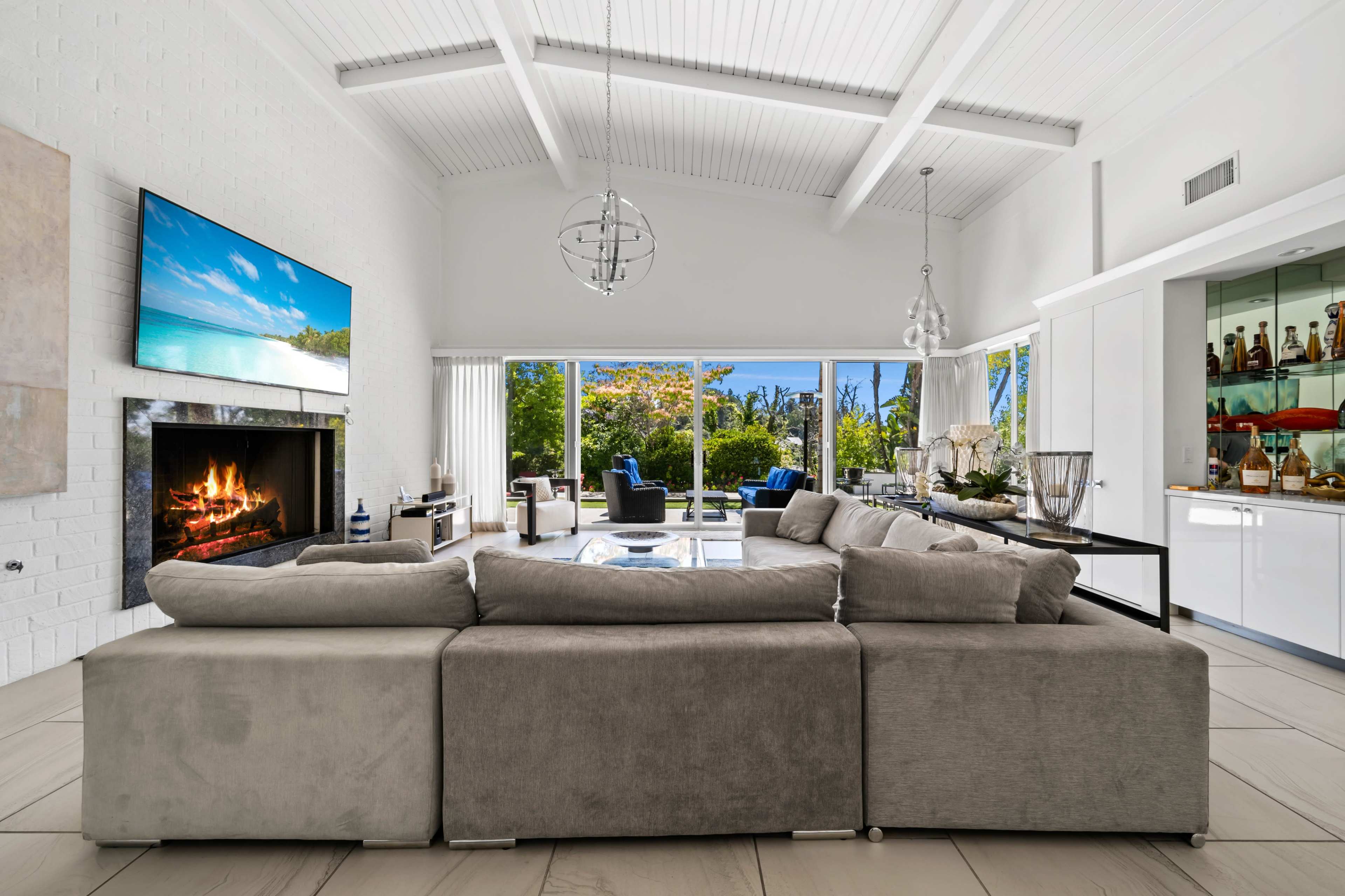 The image shows a modern living room with a large sectional sofa facing a television mounted on the wall, a fireplace, and large windows that provide a view of an outdoor landscape.