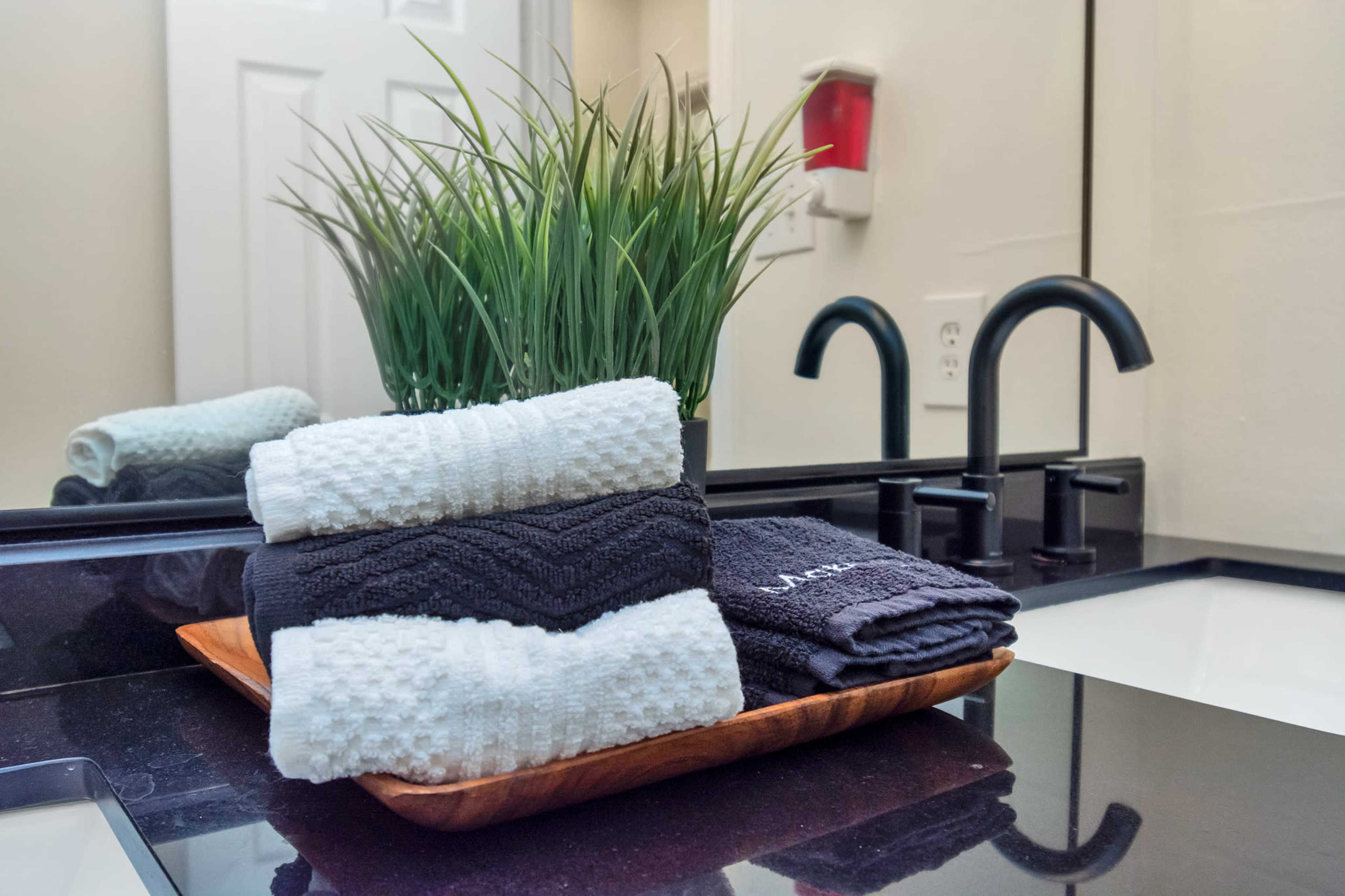 A wooden tray holds neatly stacked black and white towels next to a small potted plant in a bathroom setting with a sink and faucet visible in the background.