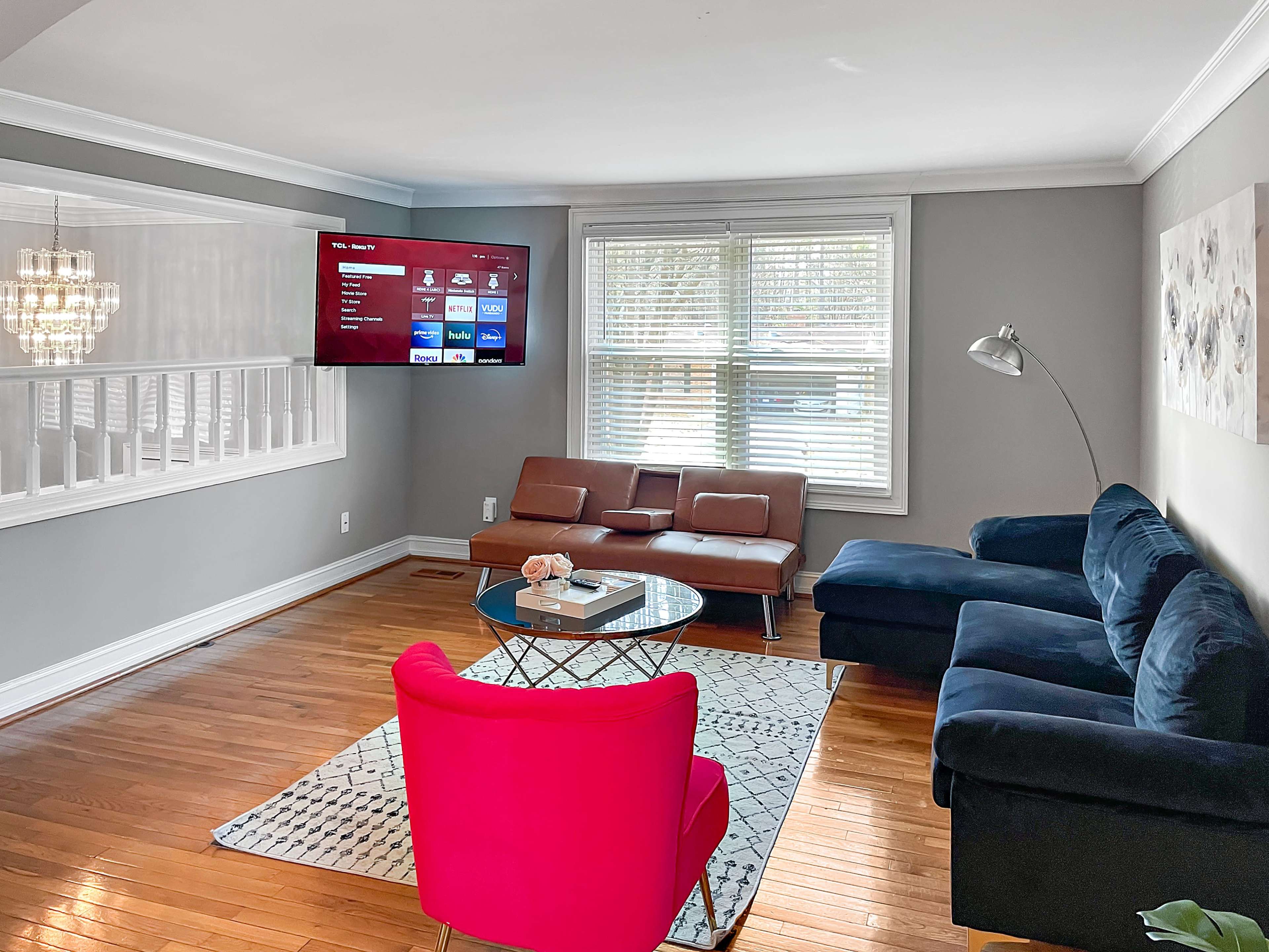 A living room featuring a dark blue couch, a brown loveseat, a pink armchair, a glass coffee table, and a wall-mounted television, all set against light gray walls and hardwood flooring.