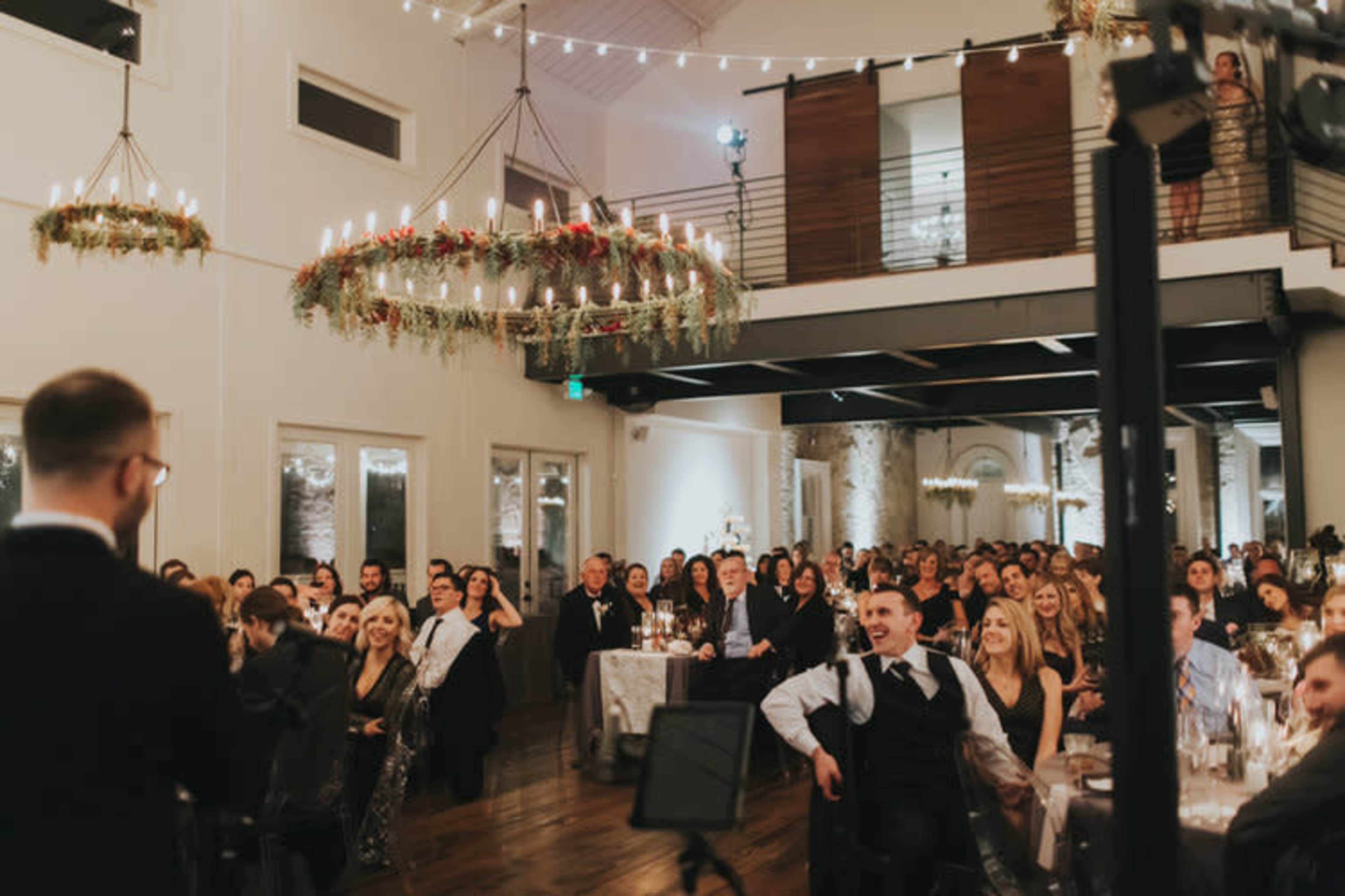 A crowd of guests dressed in formal attire attentively listens to a speaker at an event in a well-lit venue with chandeliers and elegant decor.