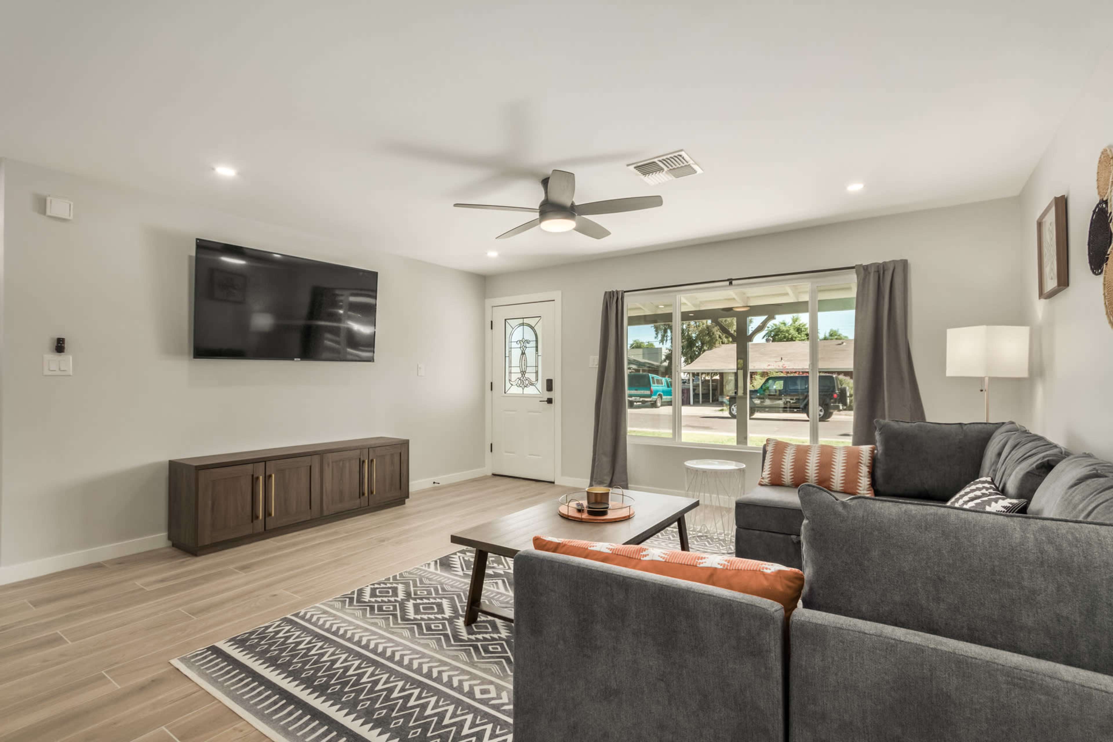 A modern living room features a gray sectional sofa, a coffee table, a wall-mounted television, and a large window with natural light pouring in.