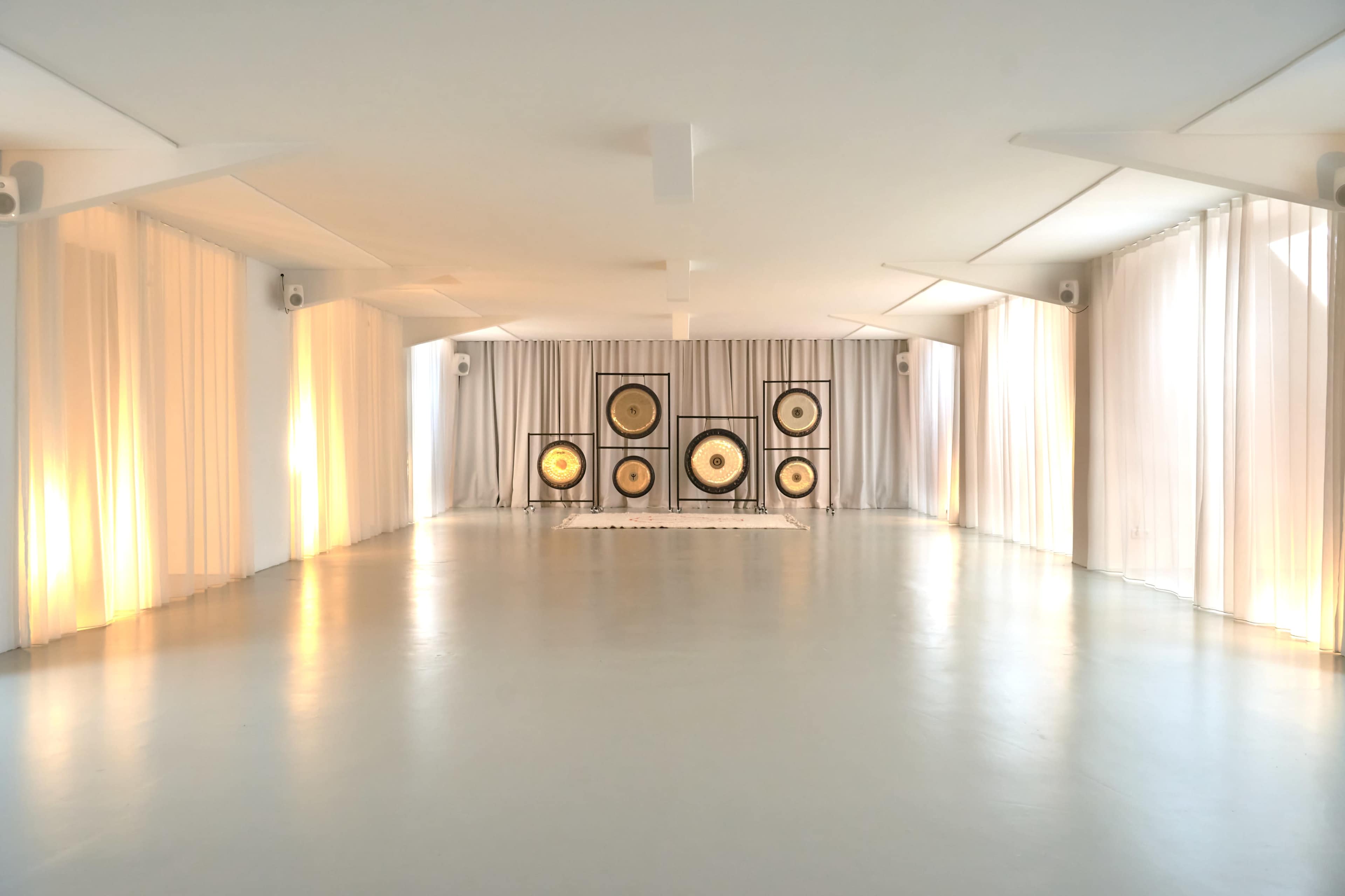 The image shows a minimalist interior space featuring large gongs arranged against a backdrop of white curtains and soft lighting.