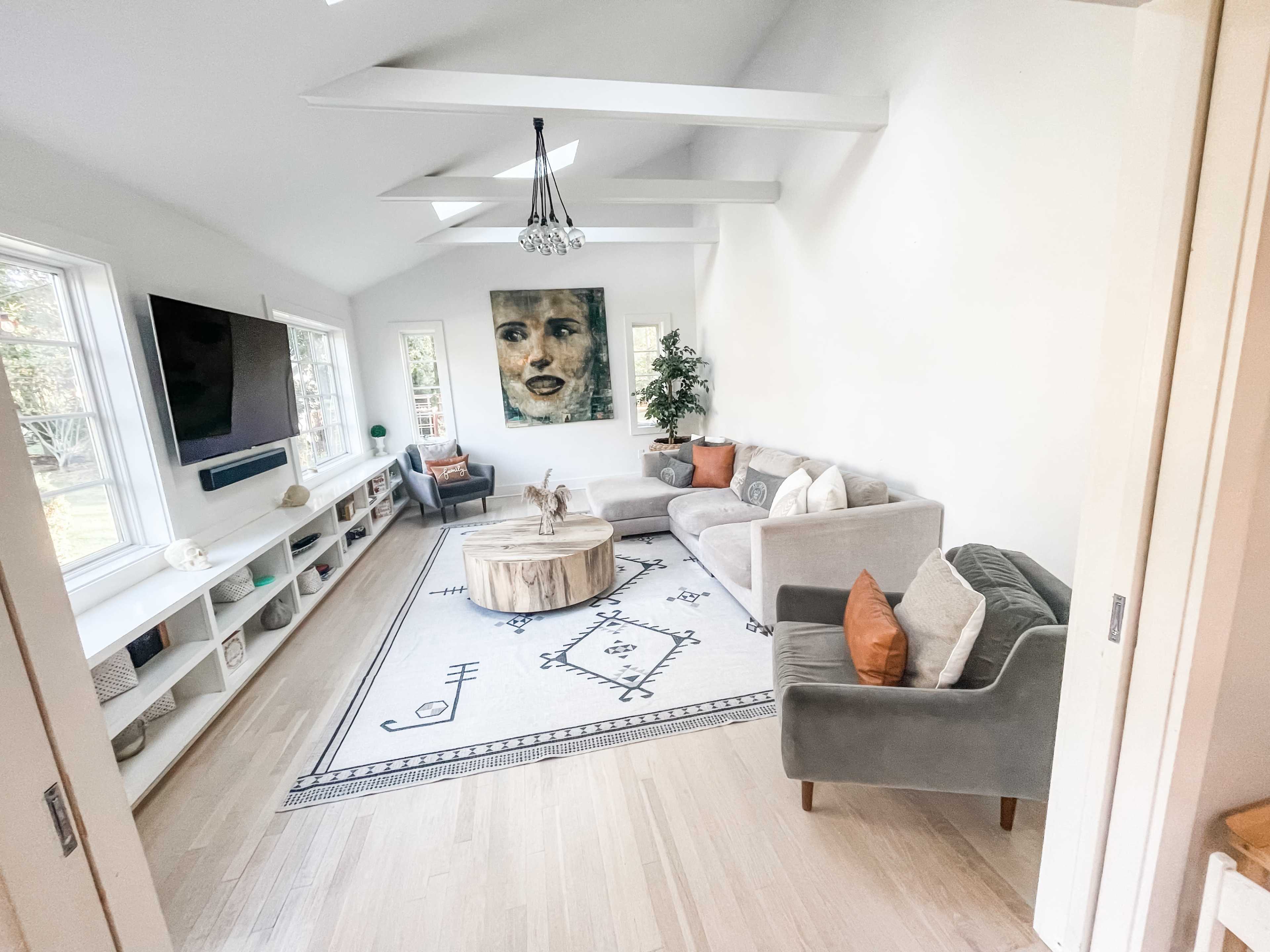 A modern living room with a gray sofa and a green armchair, a round wooden coffee table, and a large artwork on the wall, with a light-colored rug and a television mounted on a white cabinet.