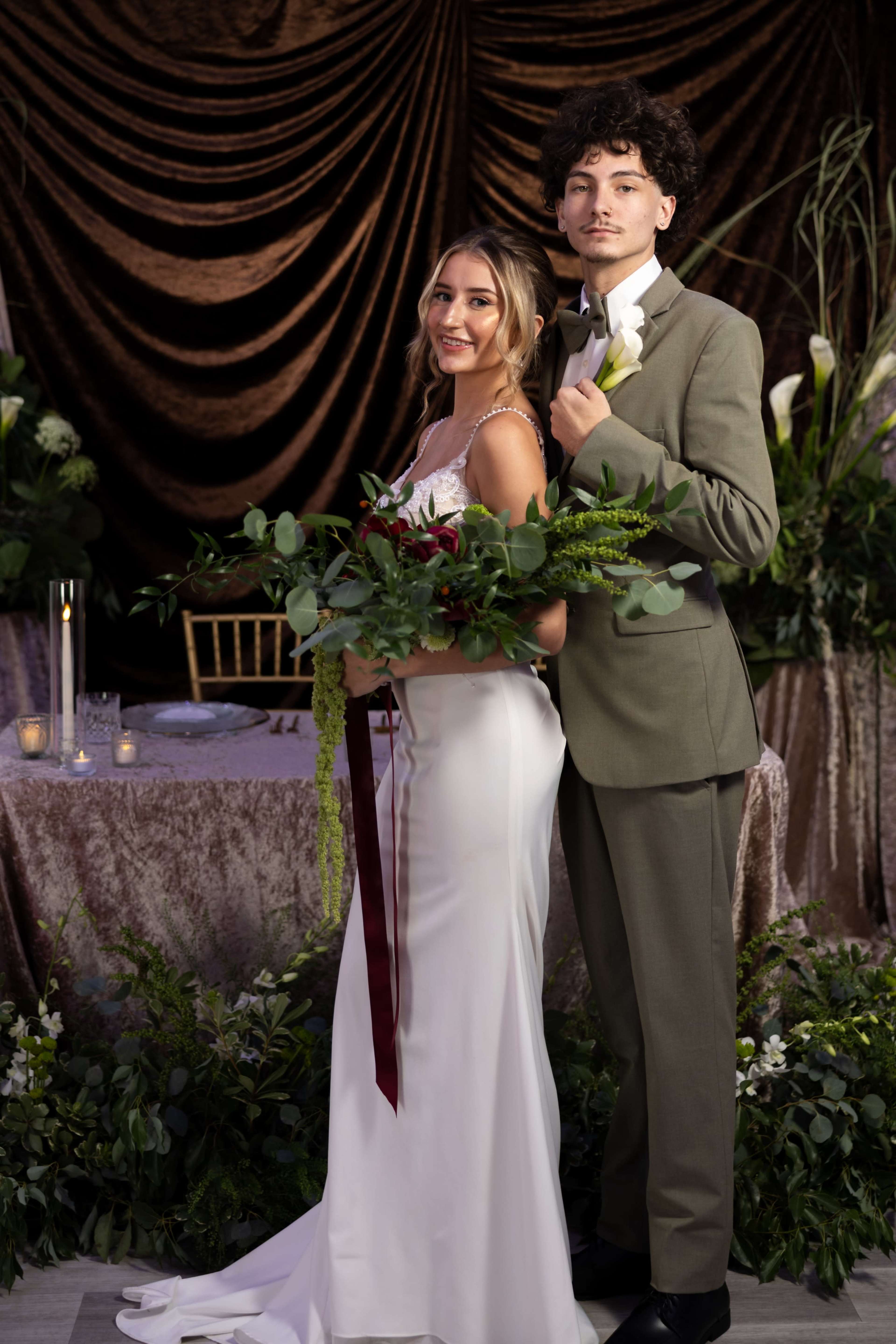 A couple stands together in formal attire, with the woman holding a bouquet and the man wearing a suit, against a backdrop of draped fabric and greenery.