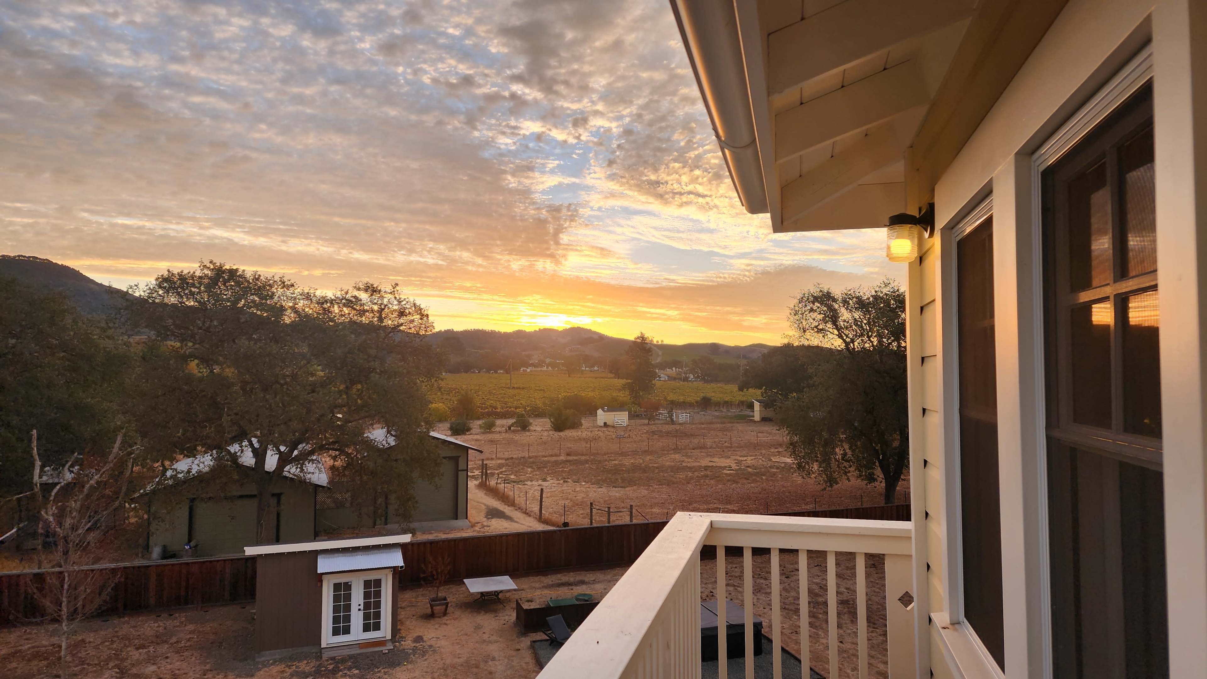 The image shows a view from a balcony overlooking a landscape with vineyards, a small shed, and a sunset sky filled with clouds.
