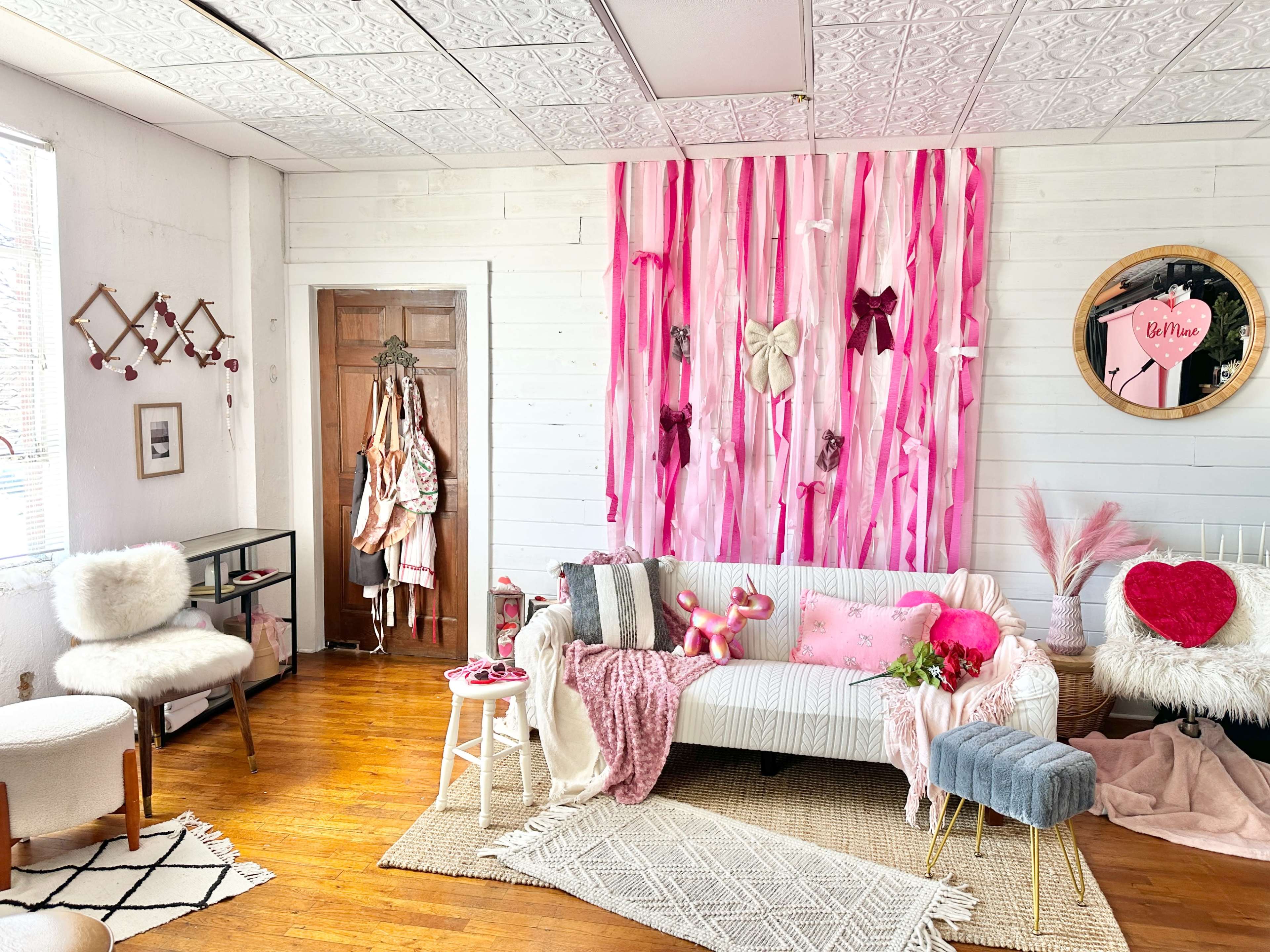 The image shows a brightly colored room decorated with pink fabrics, a white sofa, and various plush items, including a heart-shaped pillow, along with a door and wall adorned with ribbons and bows.