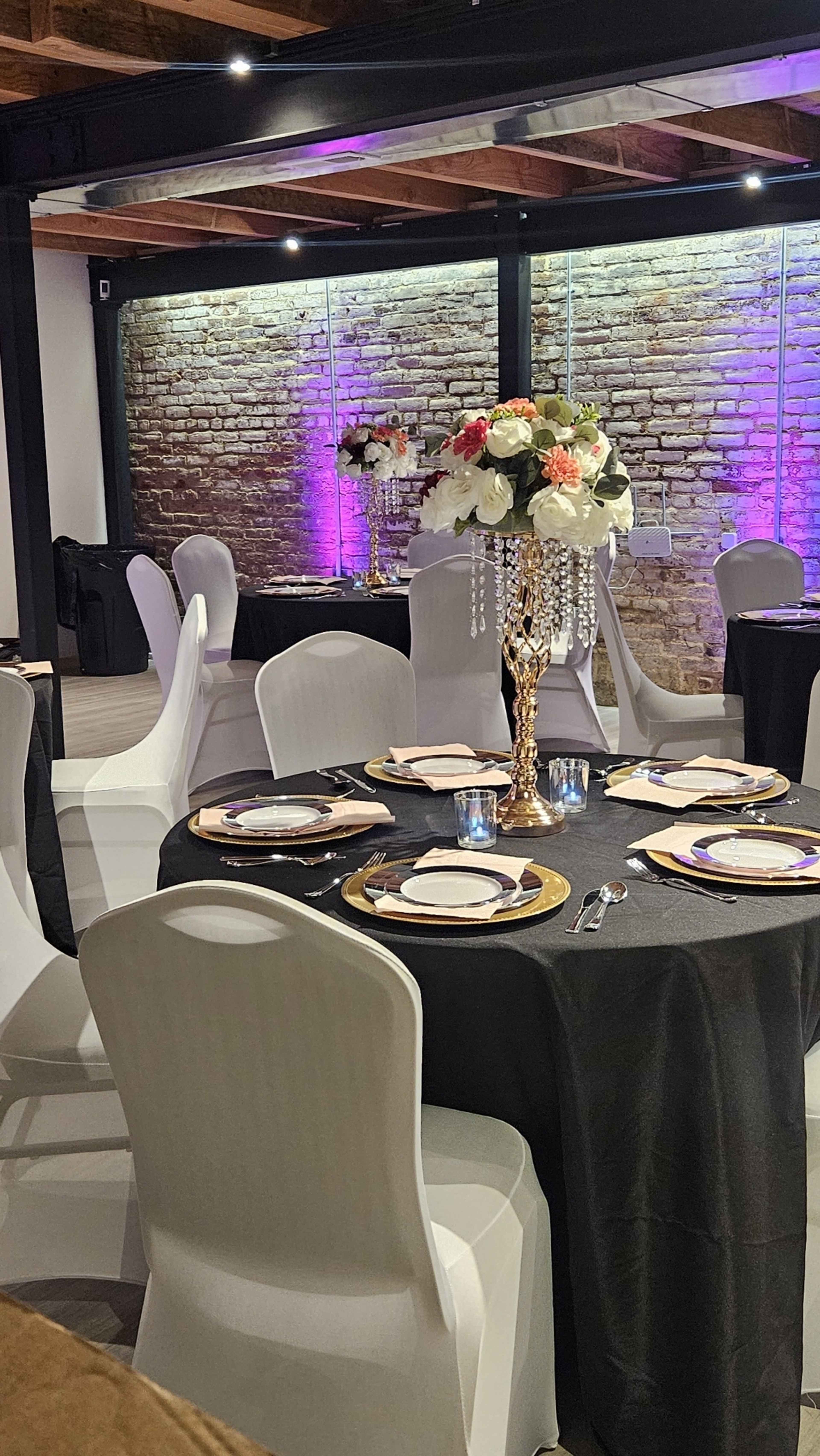 A modern dining setup features elegantly arranged tables with black tablecloths, decorative centerpieces, and purple lighting against a brick wall.