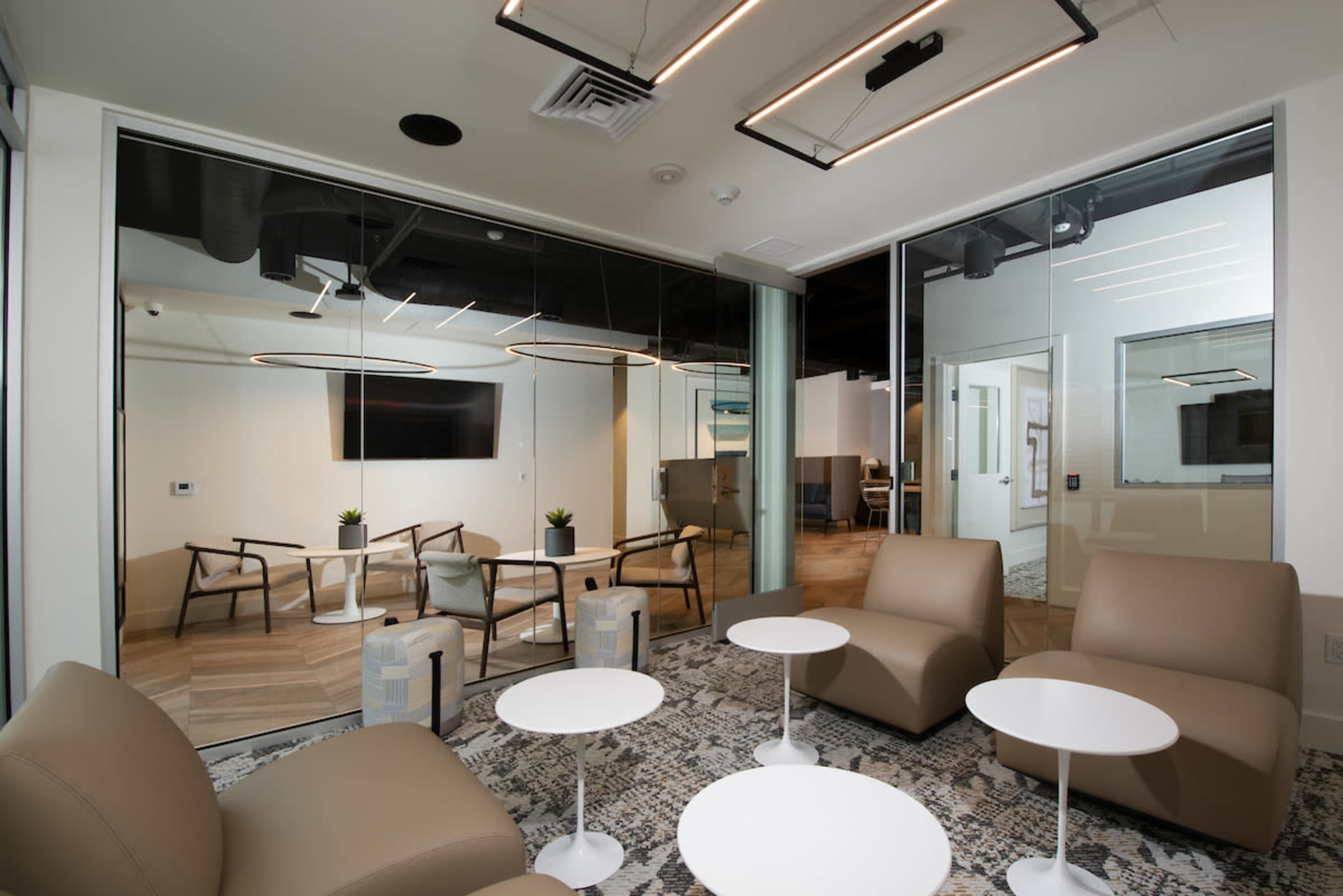 A modern office lounge area with curved seating, round white tables, and glass-walled conference rooms in the background.