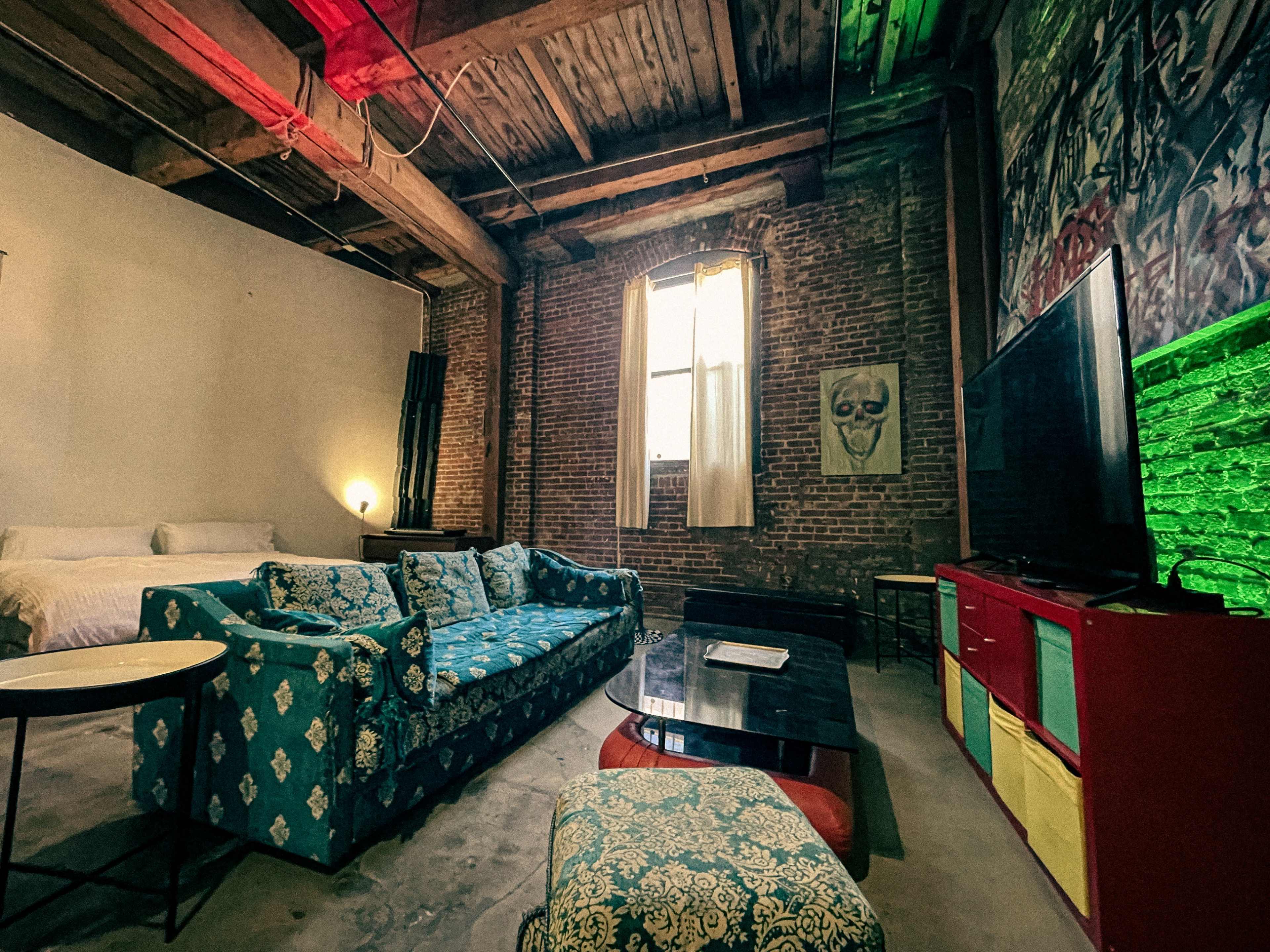 A spacious loft-style living area featuring exposed brick walls, a colorful couch, a coffee table, and a large window with sheer curtains.