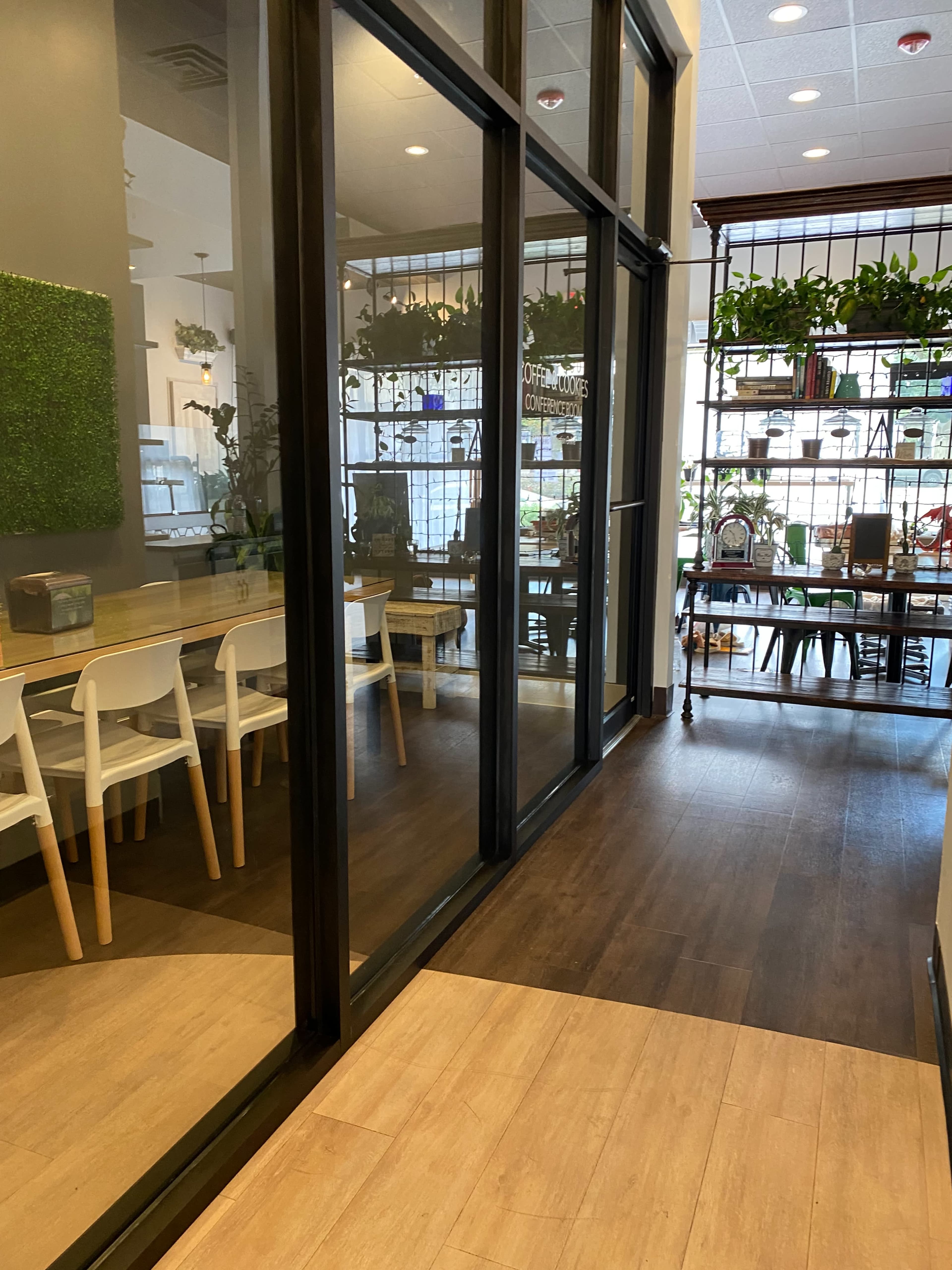 A modern interior space with glass walls, featuring a wooden floor, white chairs along a table, and numerous plants in a well-lit environment.