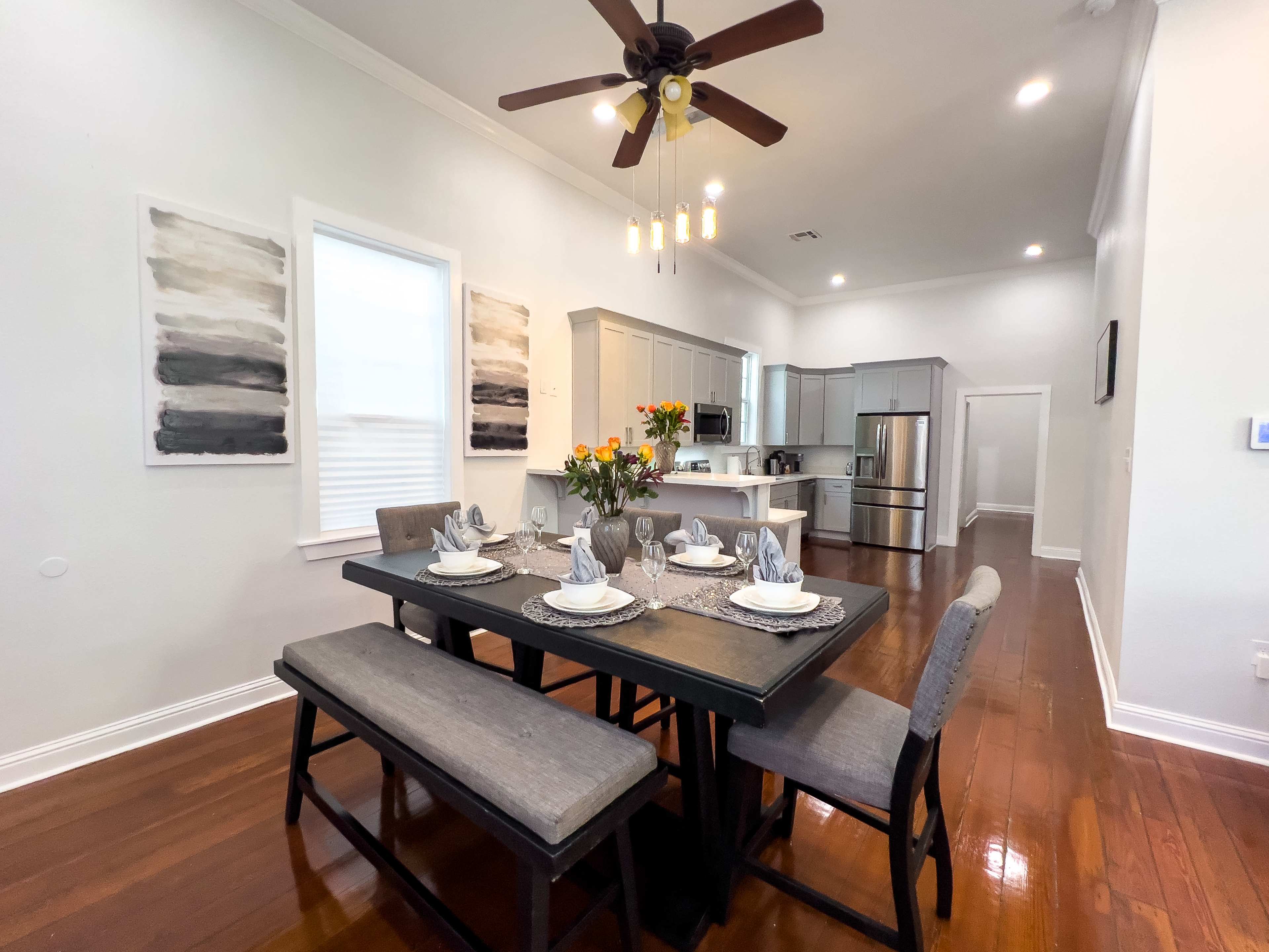 A dining area features a dark wooden table set with plates and utensils, surrounded by benches and chairs, with a kitchen visible in the background.