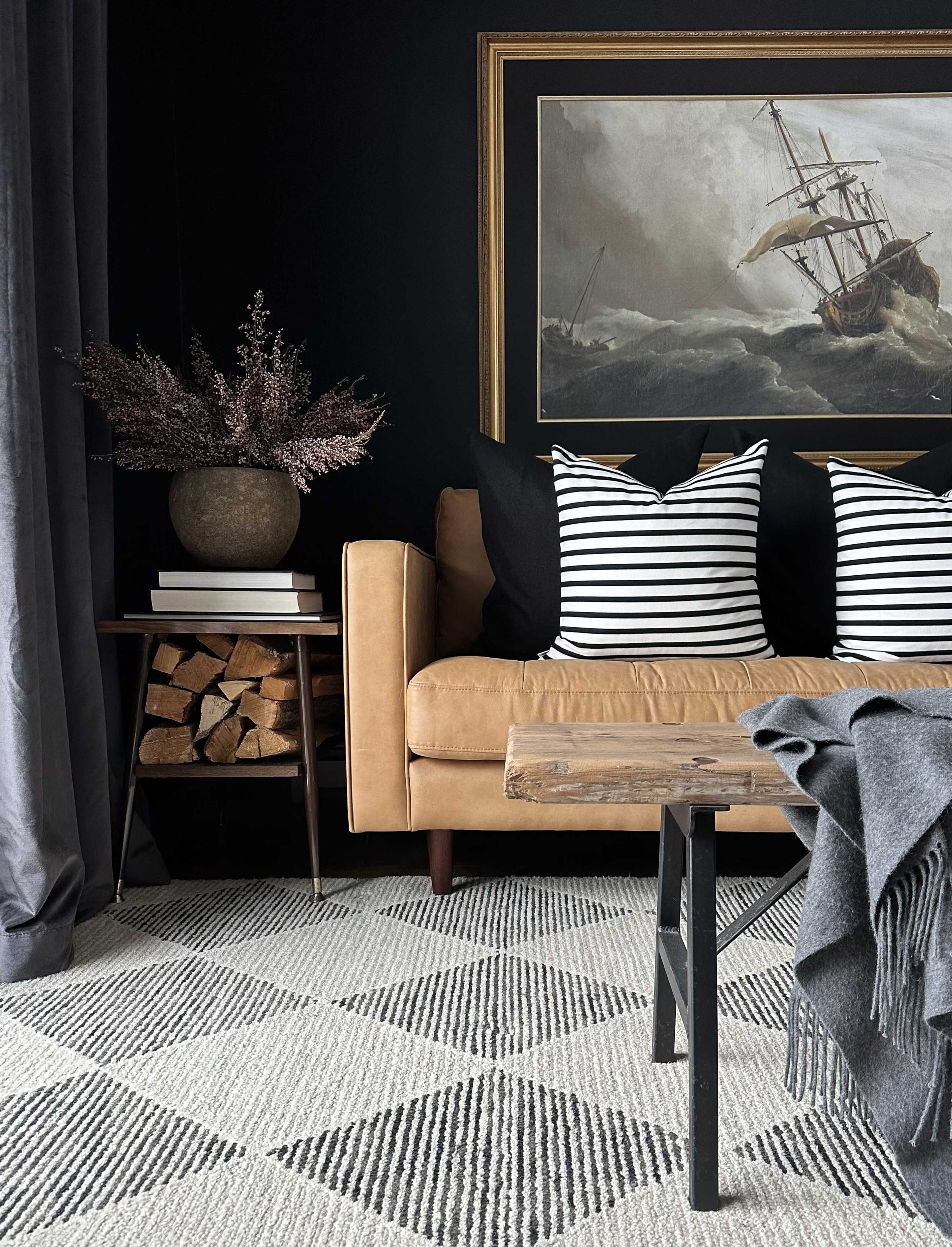 A tan leather sofa is positioned against a dark wall, complemented by striped pillows, a wooden coffee table, and a stack of firewood beside a dark vase with decorative flowers.
