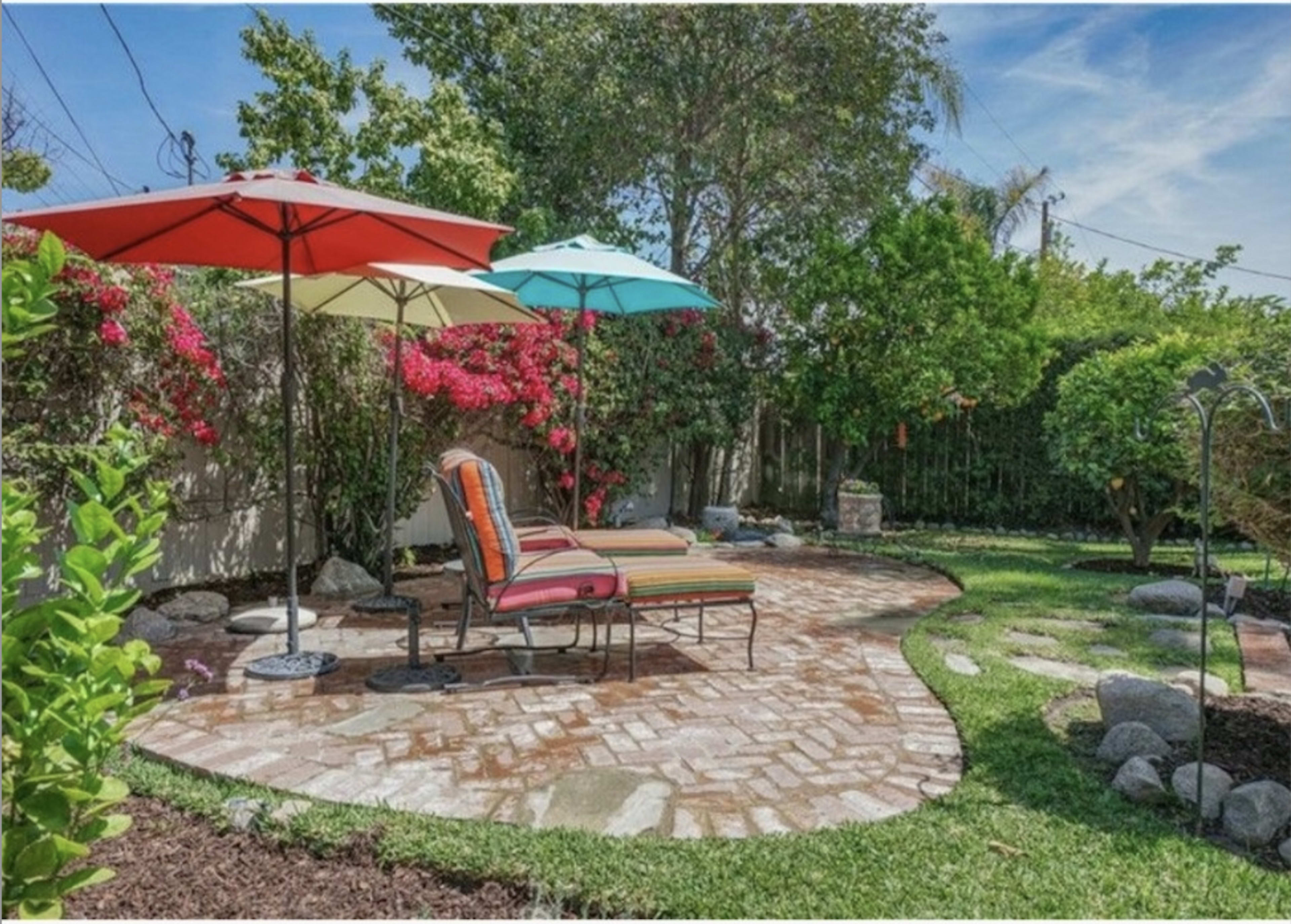 The image shows a landscaped backyard with a brick pathway, two colorful umbrellas, and lounge chairs surrounded by greenery and flowering plants.
