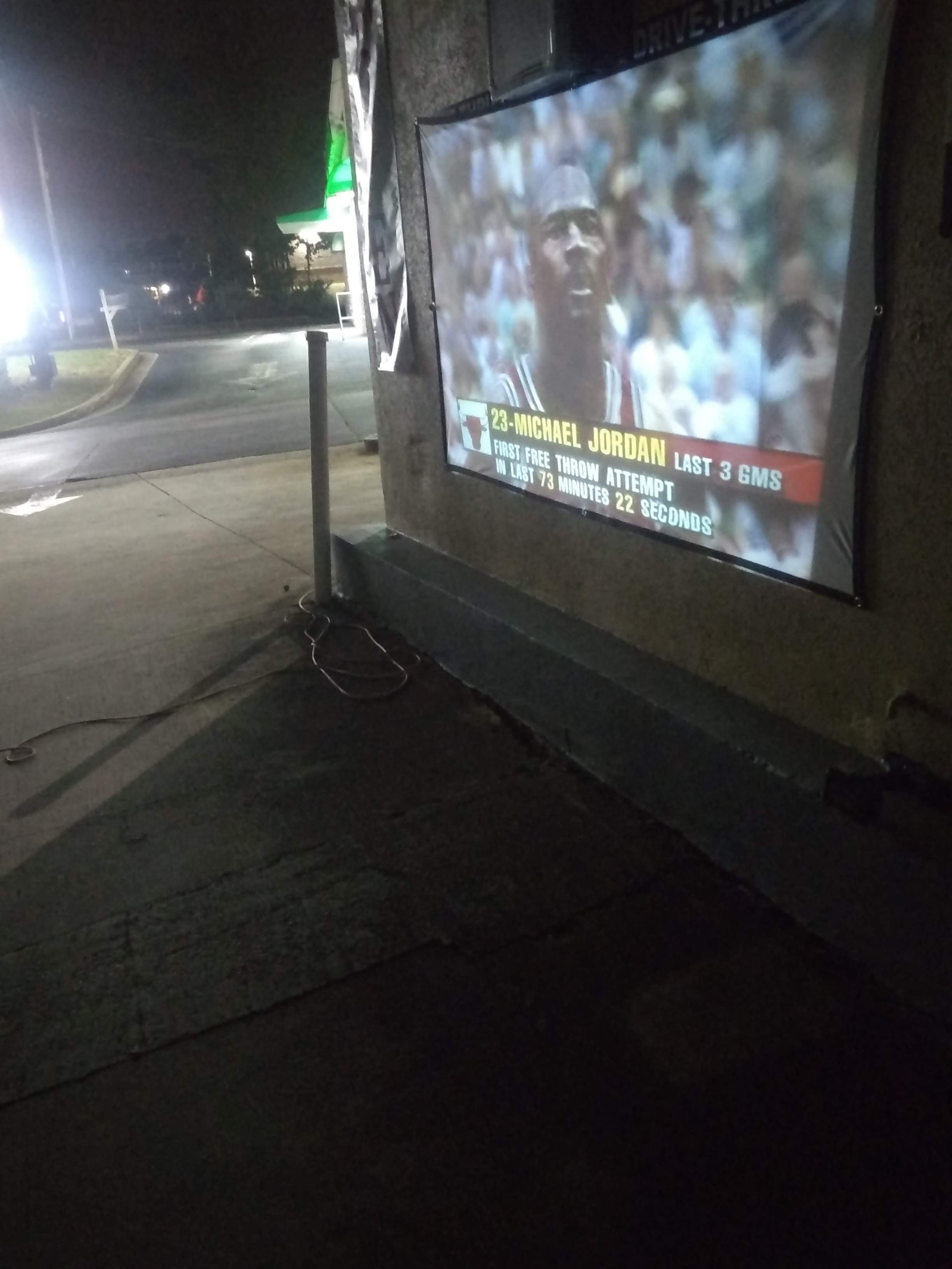 A projector displays a sports broadcast of Michael Jordan shooting a free throw, illuminating a wall in a dimly lit area.