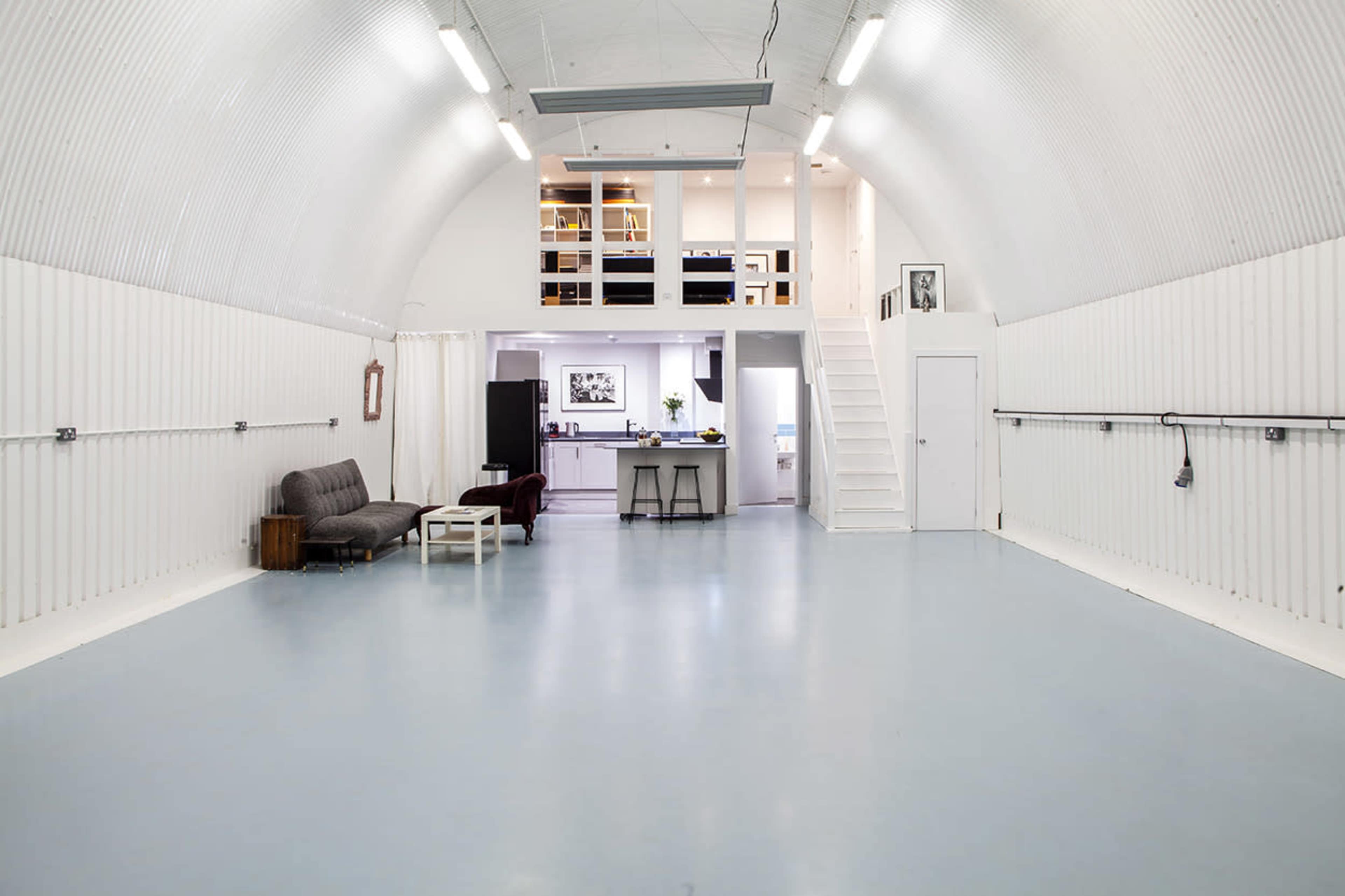 The image shows a spacious, modern interior with high arching white walls, a light blue floor, and a loft area featuring a kitchen and dining space.