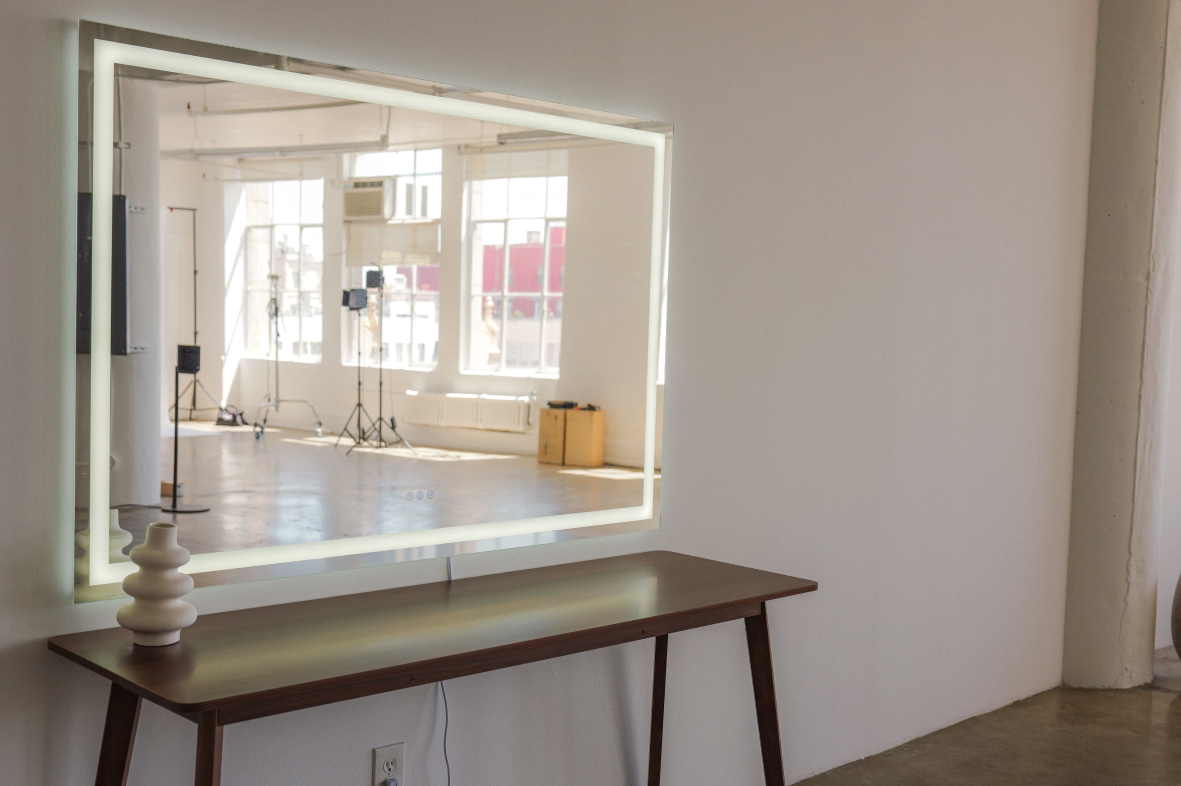 A backlit mirror is mounted on a white wall above a wooden table in an empty room with large windows.