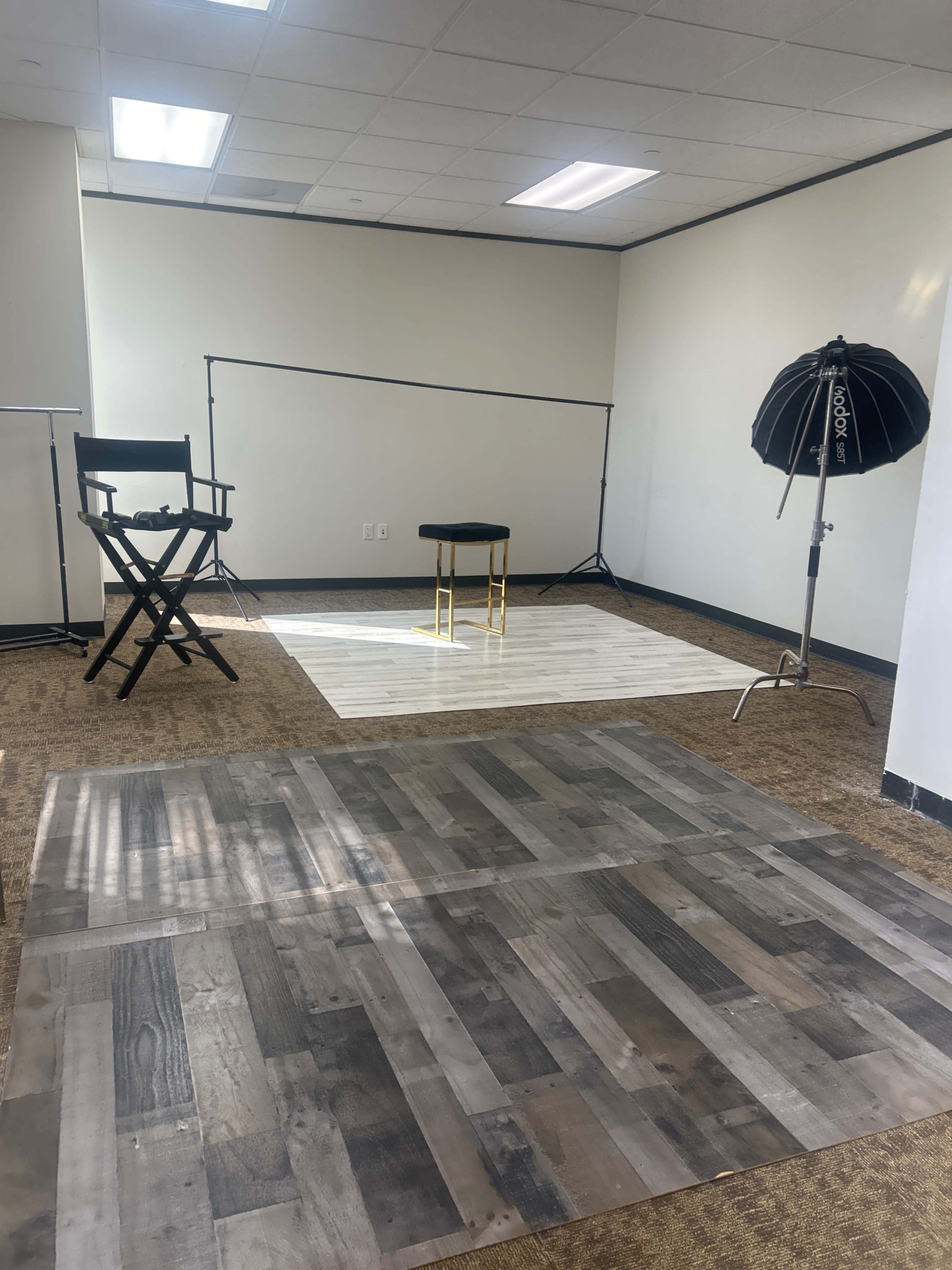The image shows a photography studio setup featuring a wooden floor platform, a director's chair, a stool, and a large umbrella light.