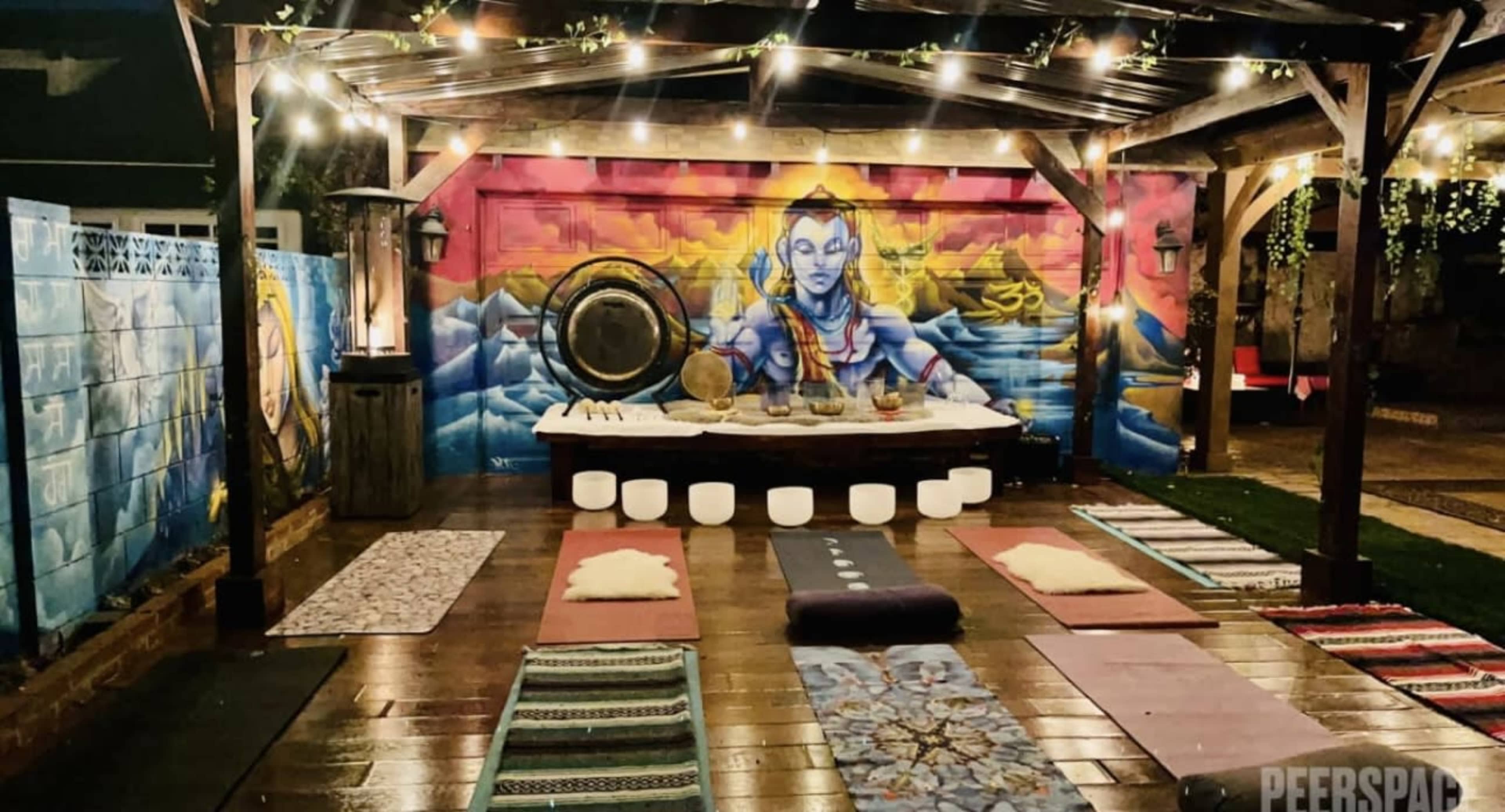The scene features an outdoor meditation space with yoga mats arranged on a wooden floor under a canopy, illuminated by string lights, and adorned with a colorful mural.
