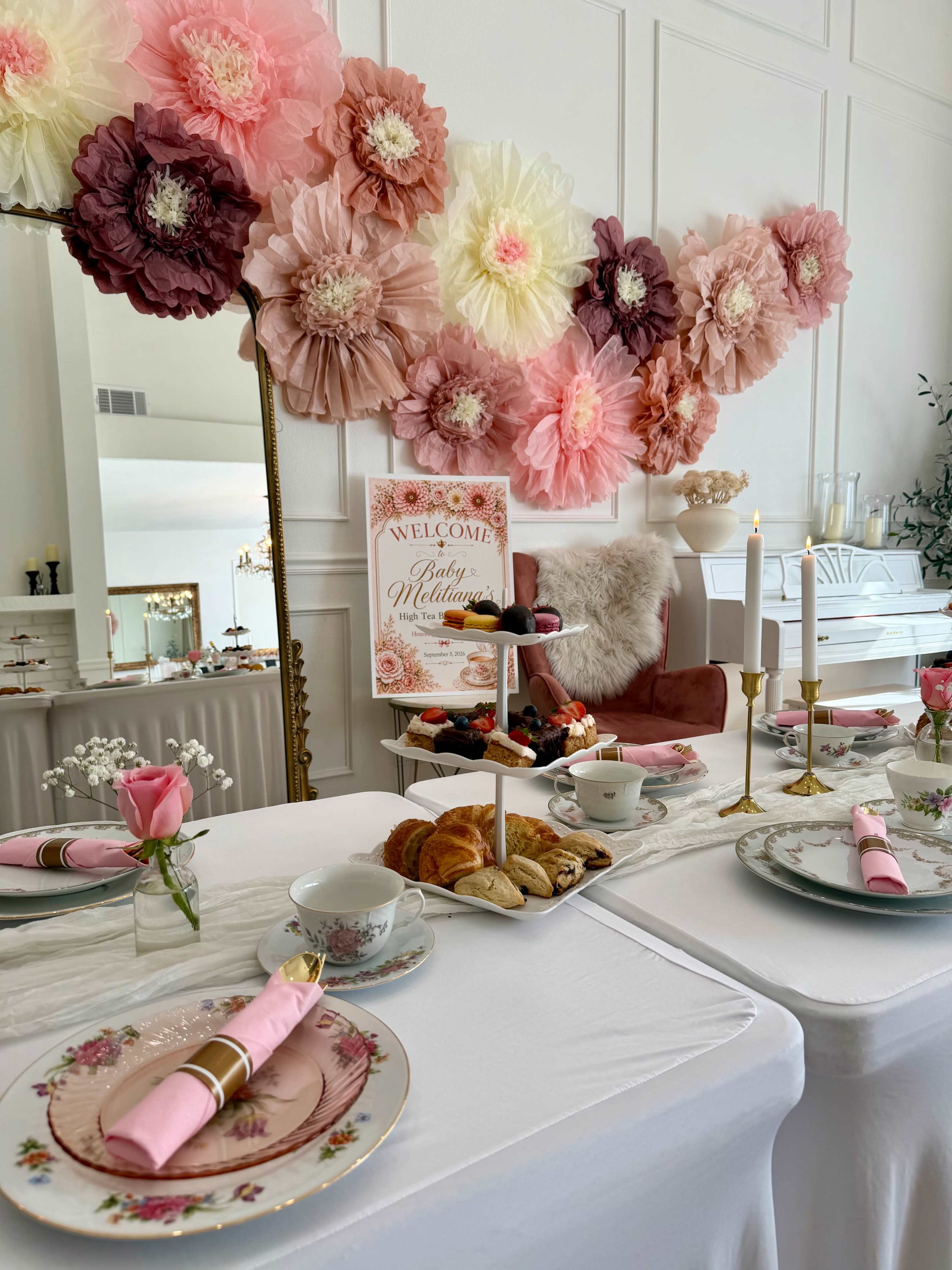 A decorative table is set for a gathering, featuring elegant tableware, pastries, and a floral backdrop.