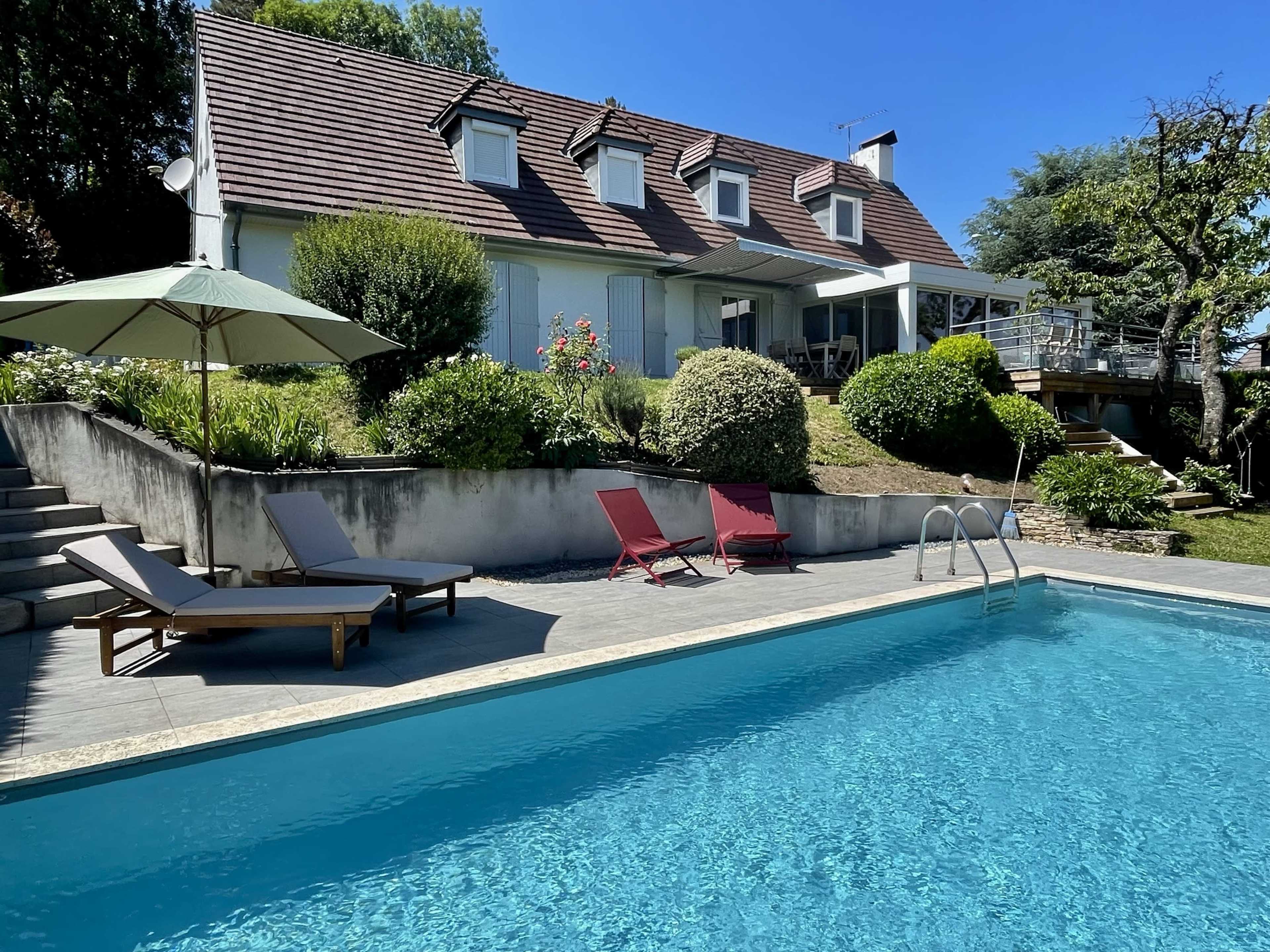 A two-story house with a sloped roof is situated behind a swimming pool, which is surrounded by lounging chairs and greenery.