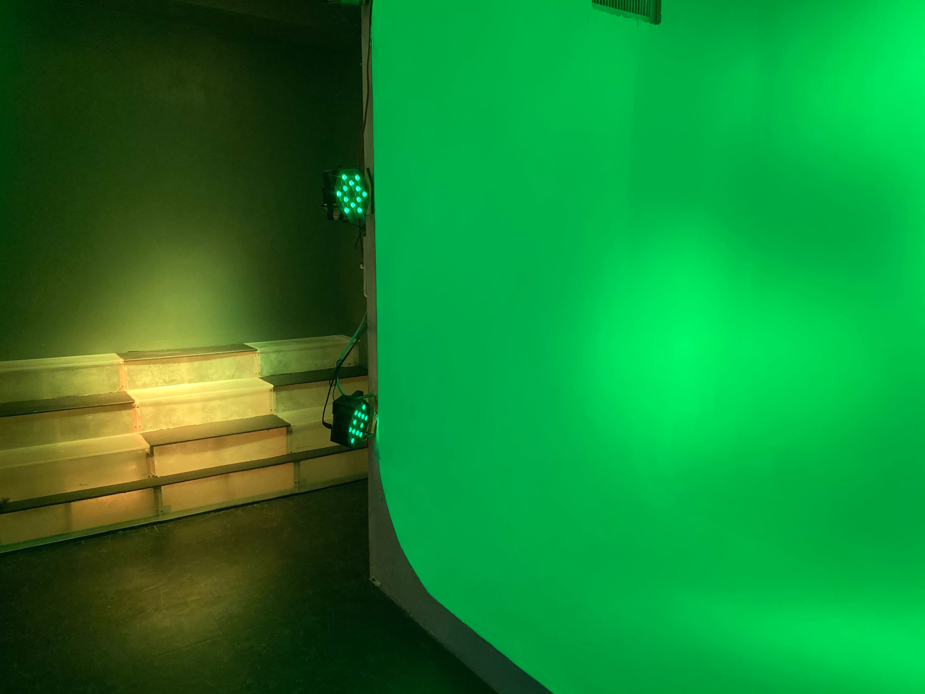 A partially lit studio with a green backdrop and two green lights illuminating the space.