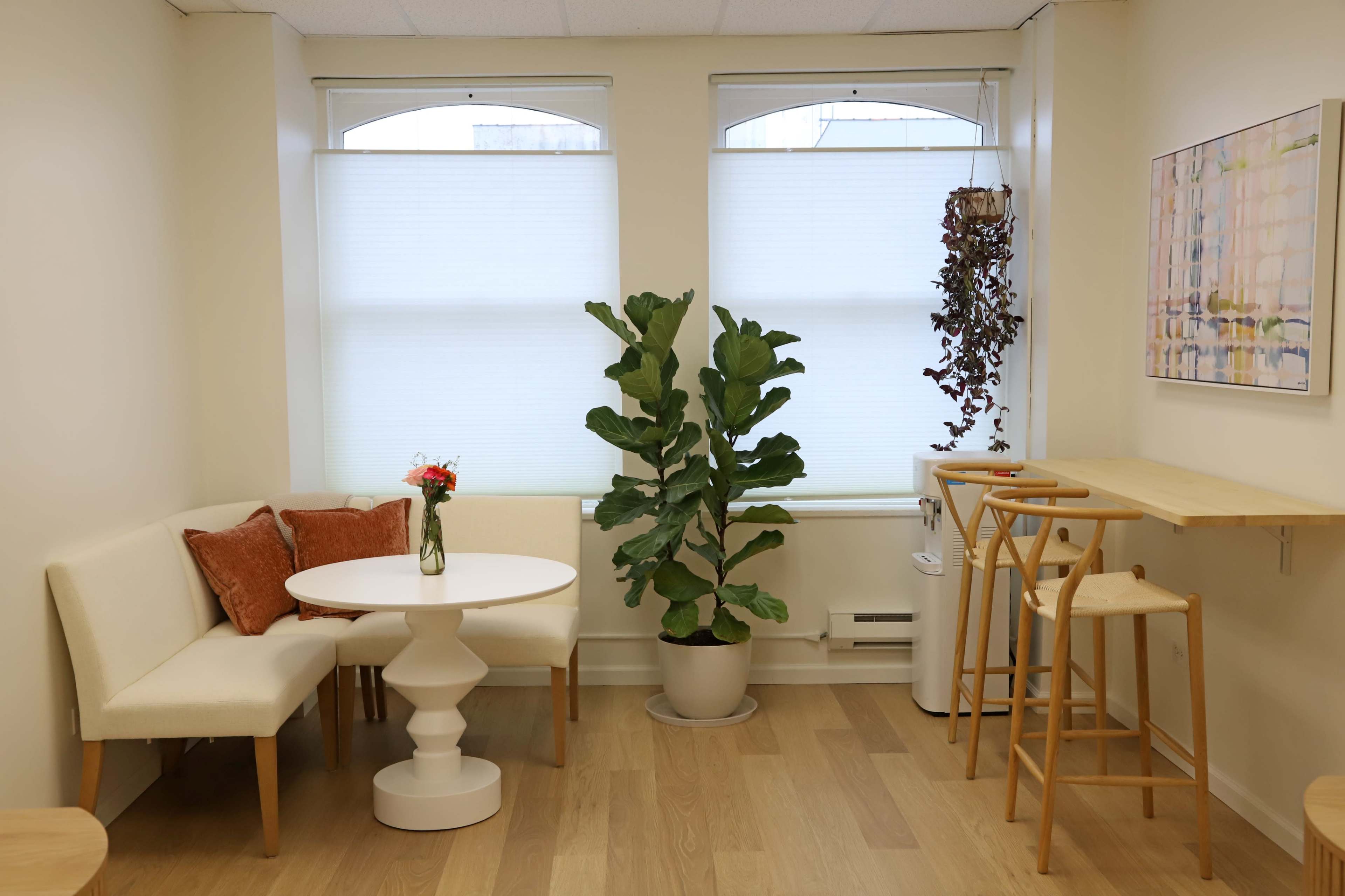 The image shows a small, bright room with a round white table surrounded by a couch and two wooden stools, adorned with a potted plant and decorative elements near the windows.
