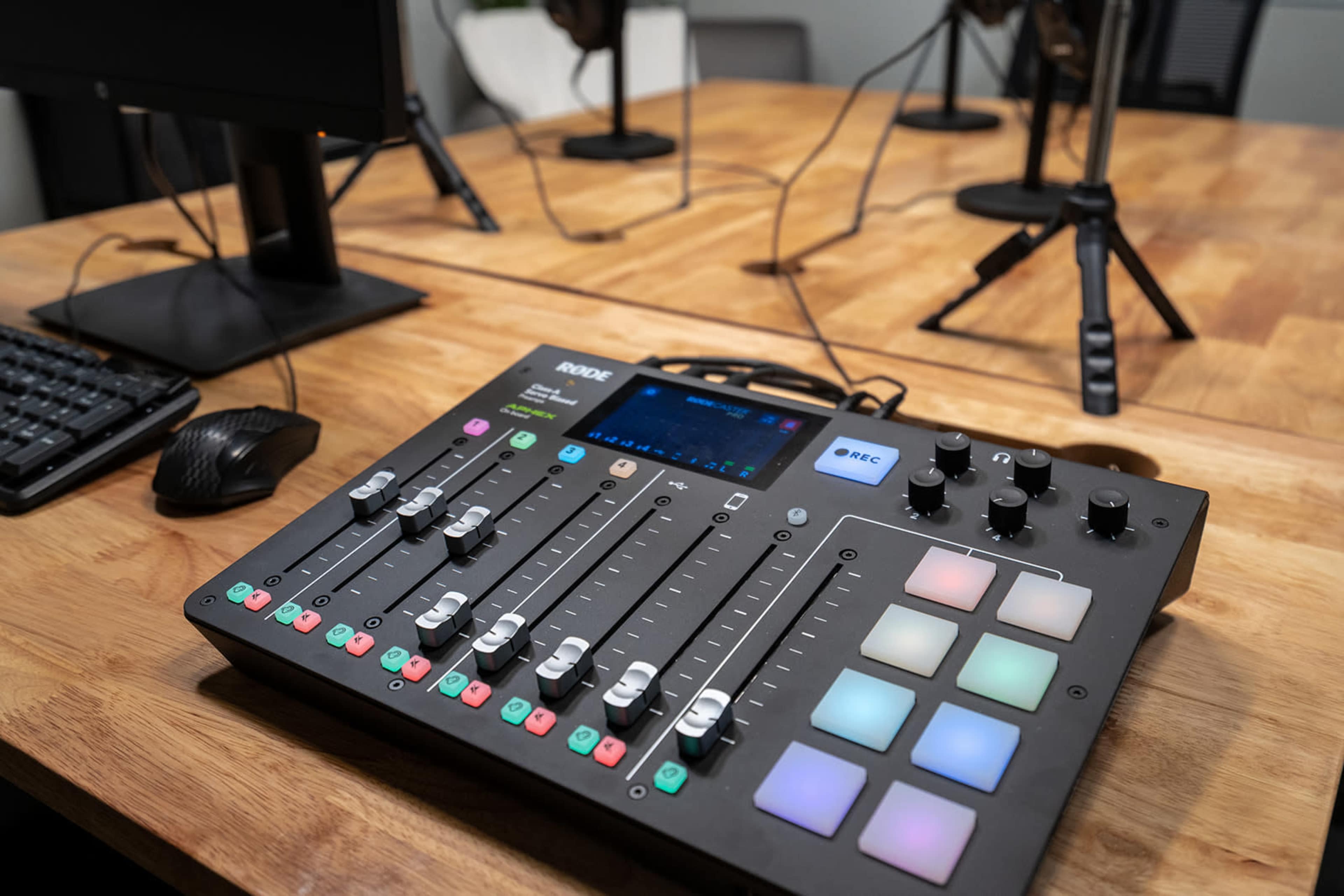 A Rode audio interface with multiple sliders and buttons sits on a wooden table in a recording studio.