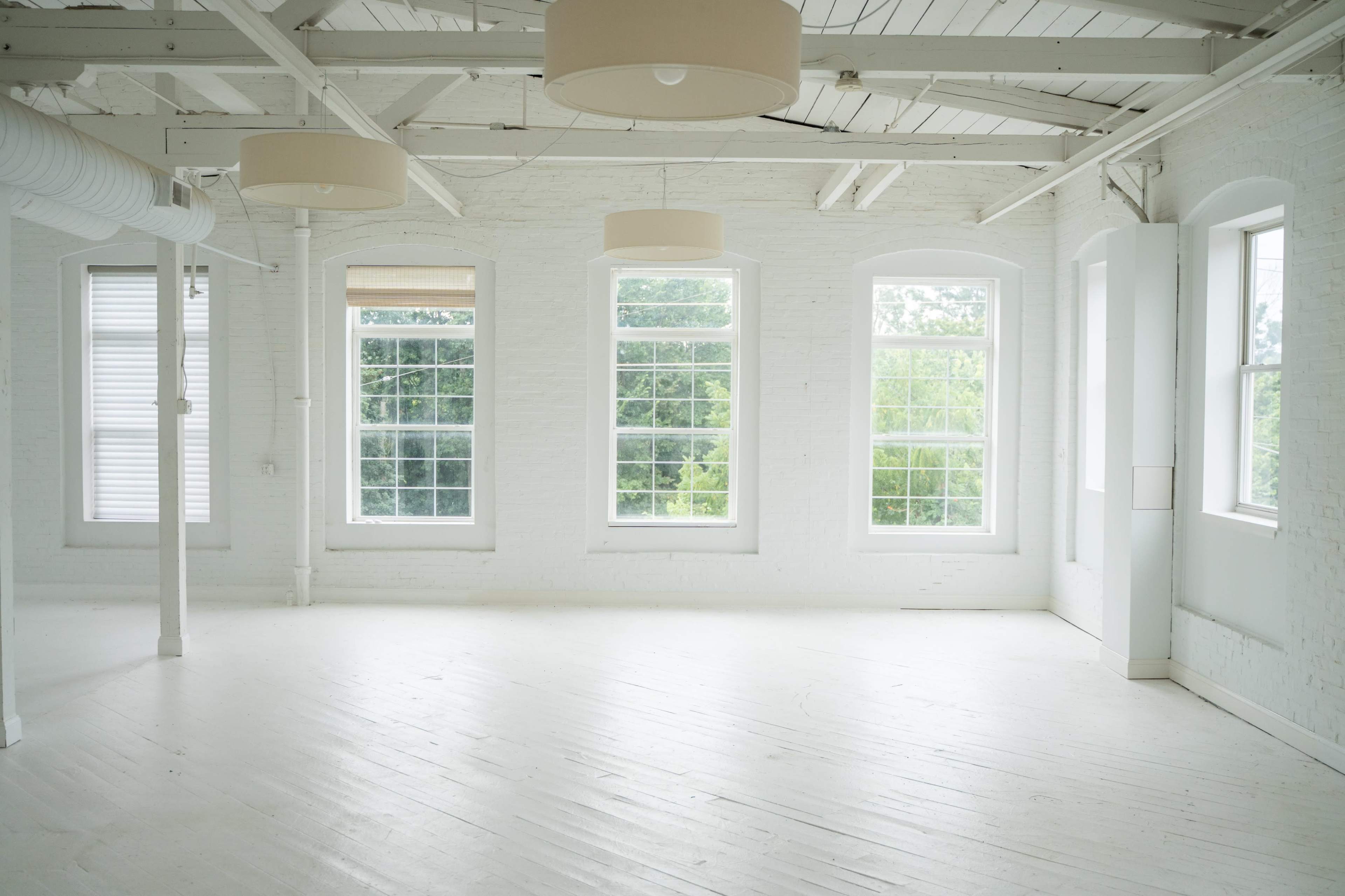 The image shows a spacious, brightly lit room with white walls, large windows, and hardwood floors.