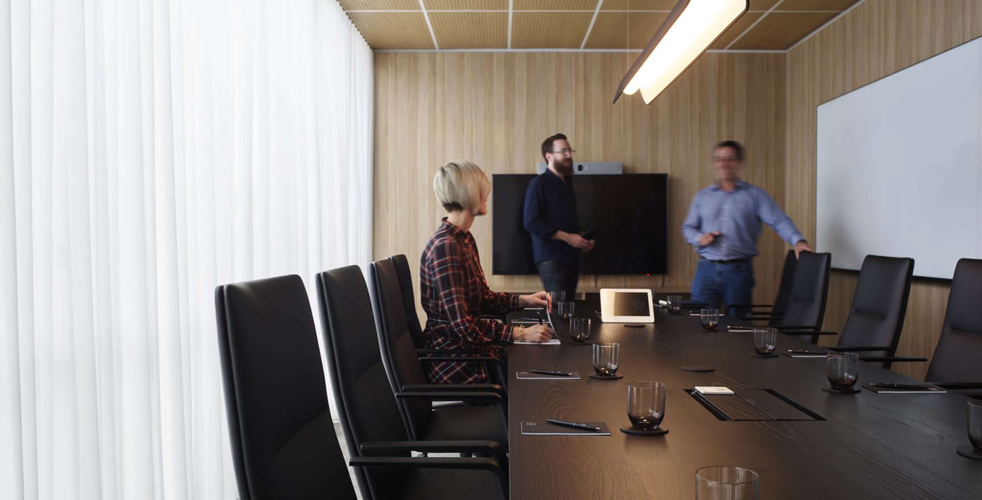 A modern conference room features a long table with black chairs, while three individuals are gathered near a presentation screen.