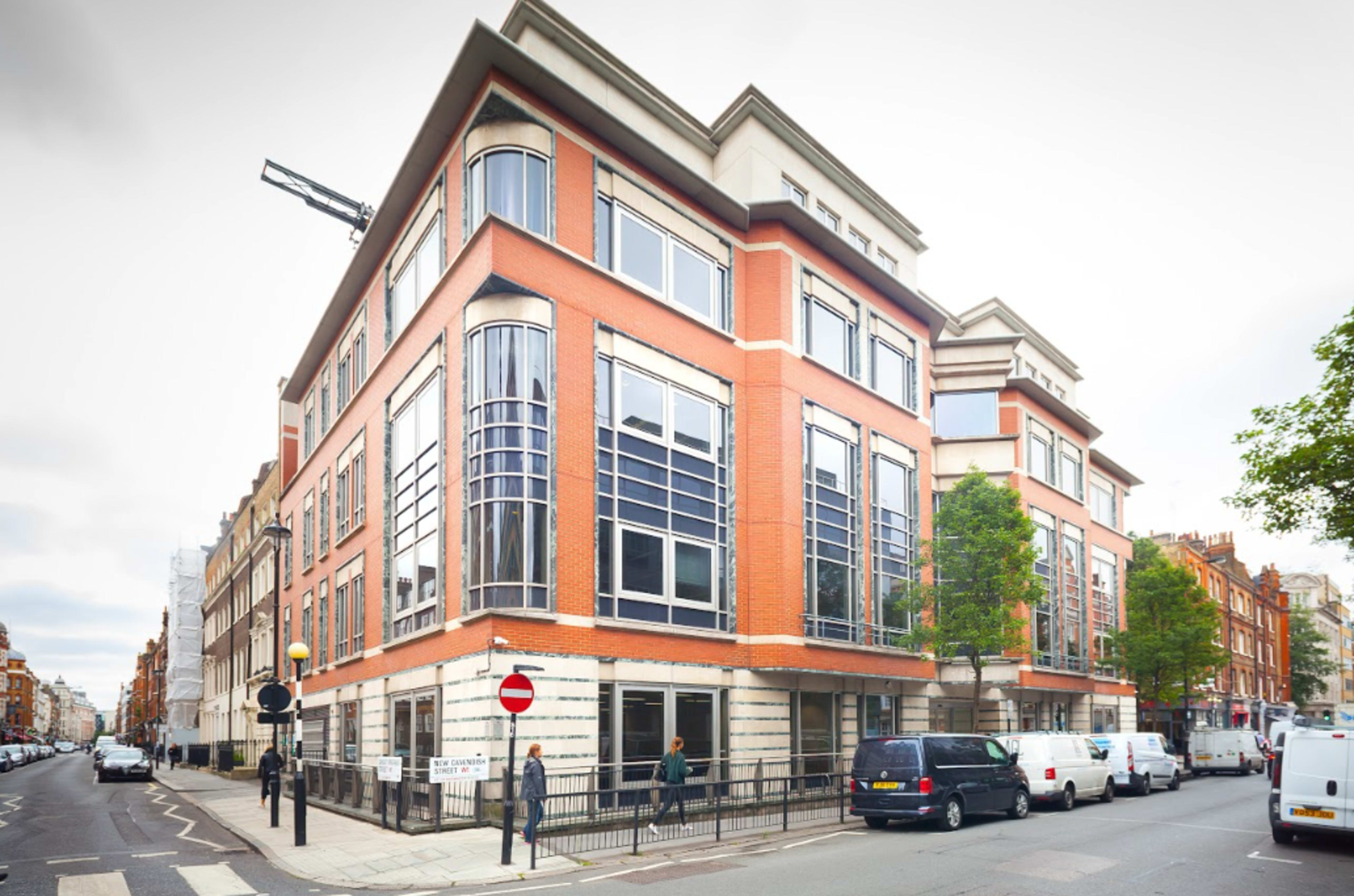 The image shows a modern red-brick building with large windows located on a street corner, surrounded by parked vehicles and greenery.