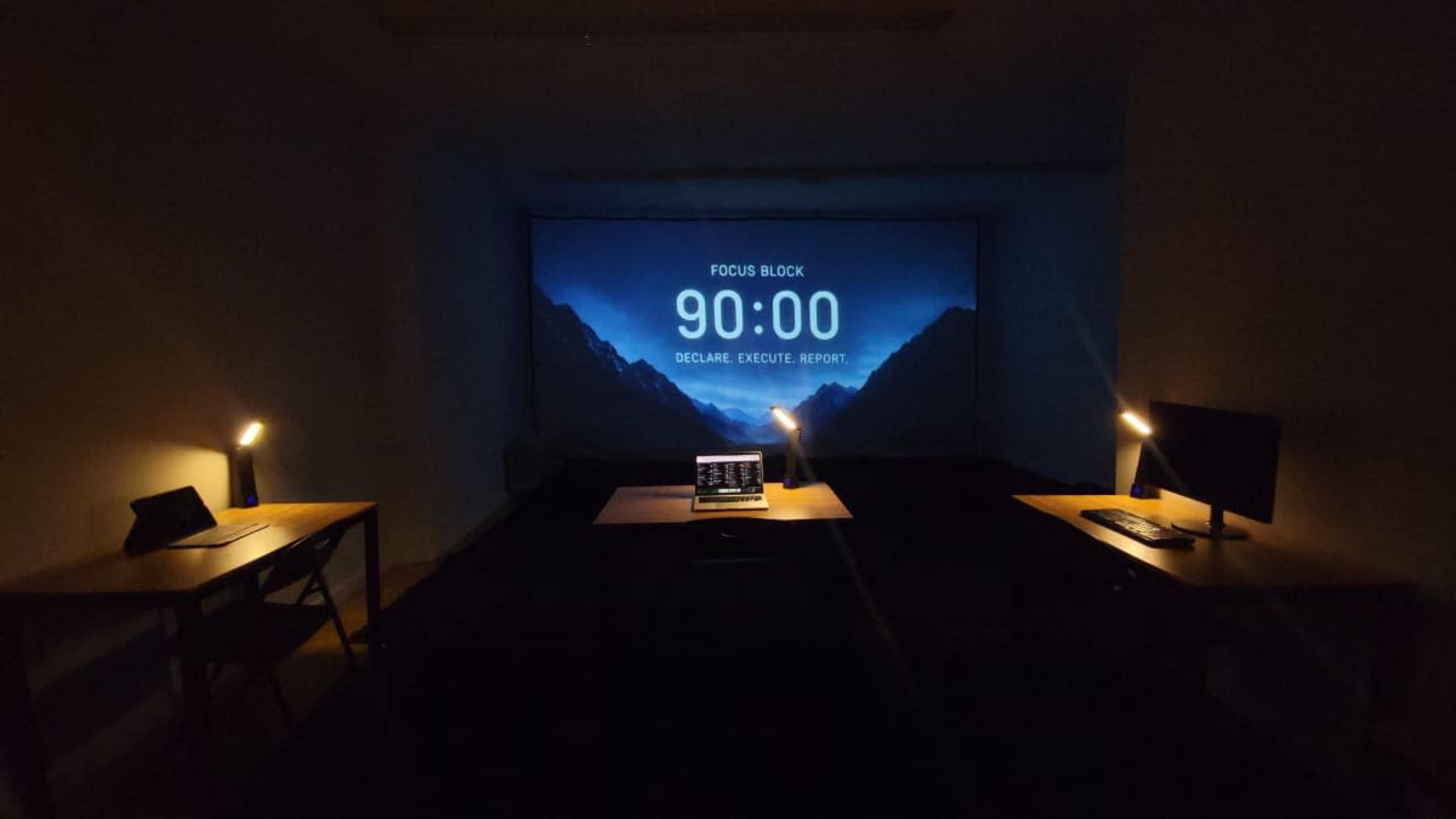 A dimly lit room features two desks with lamps and a large projection screen displaying a timer set for 90 minutes.