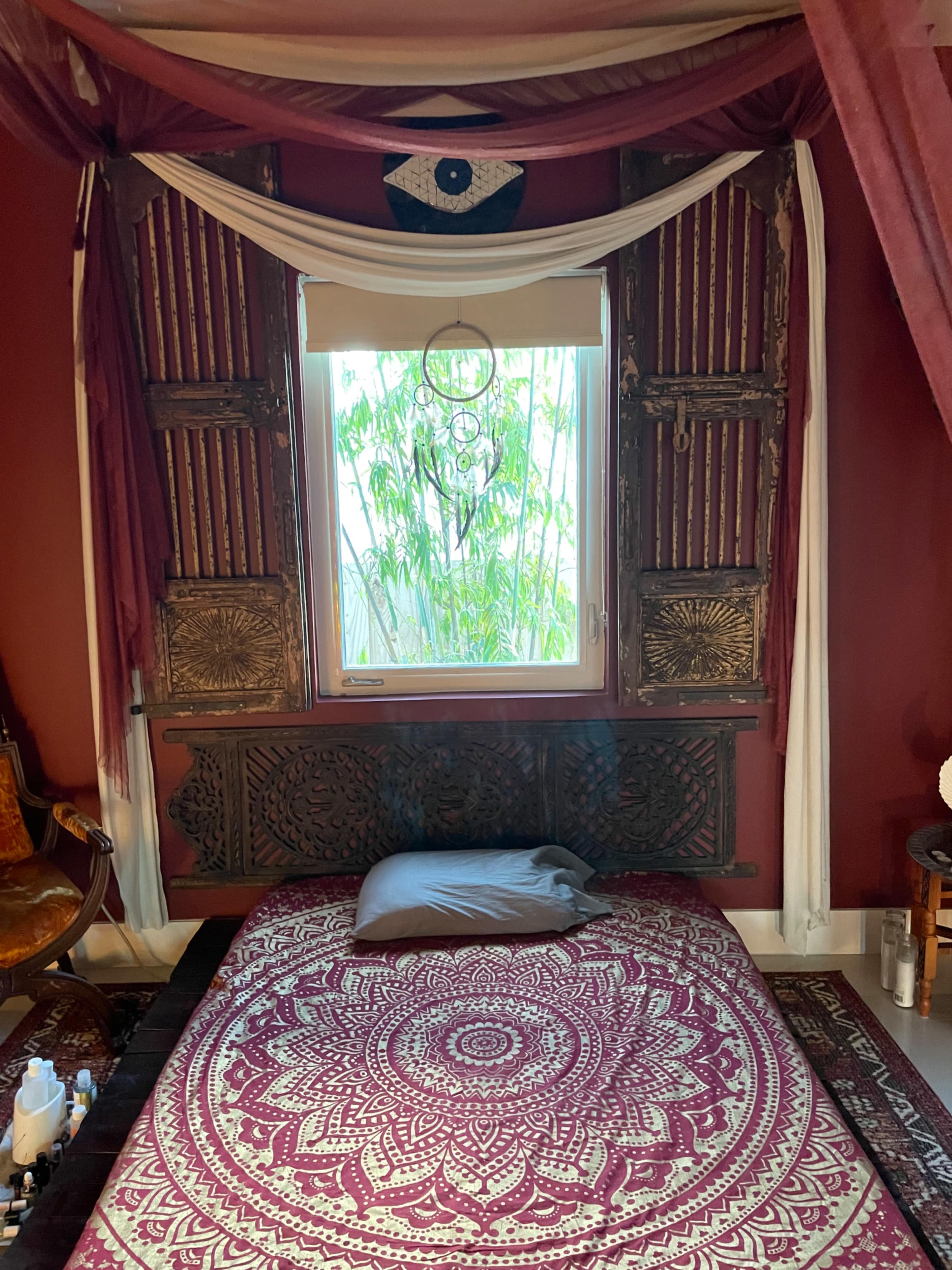 A bedroom with a low bed covered by a red patterned rug, surrounded by ornate wooden shutters and decorative fabric draping from the ceiling.