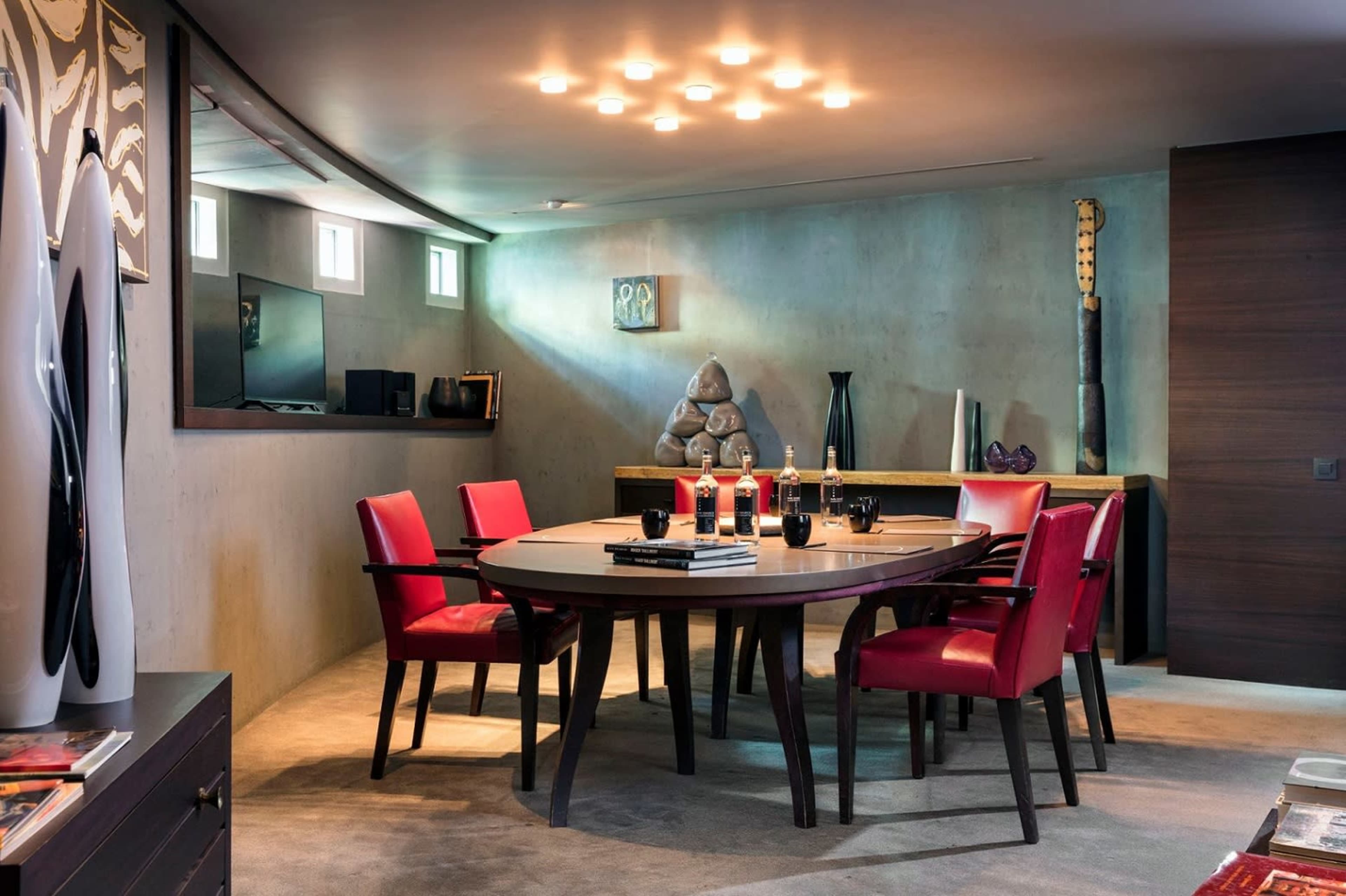 A modern dining room features a round table surrounded by red chairs, with a concrete wall and artistic decor in the background.