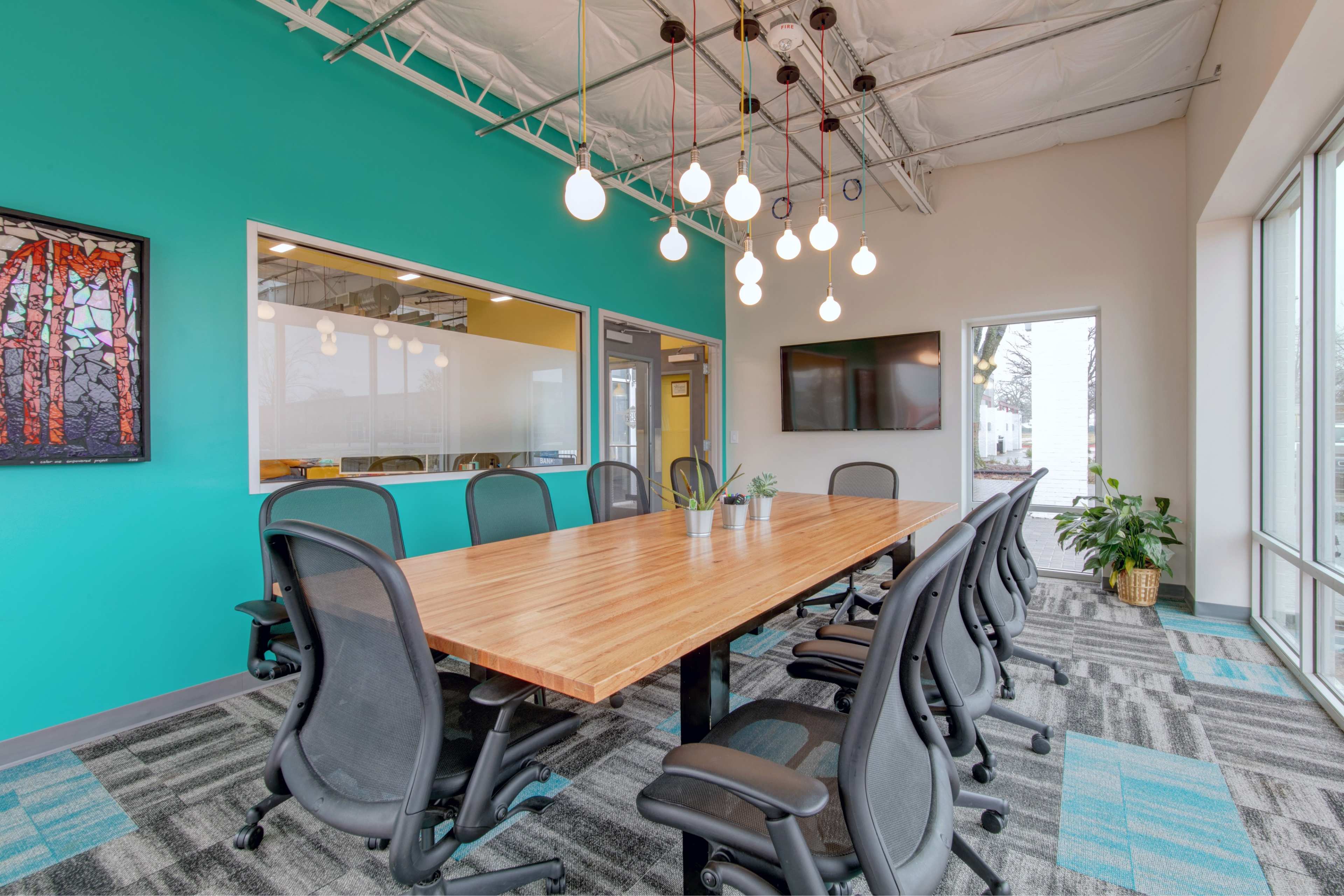 A brightly colored conference room features a large wooden table surrounded by black chairs, with modern pendant lights and large windows.