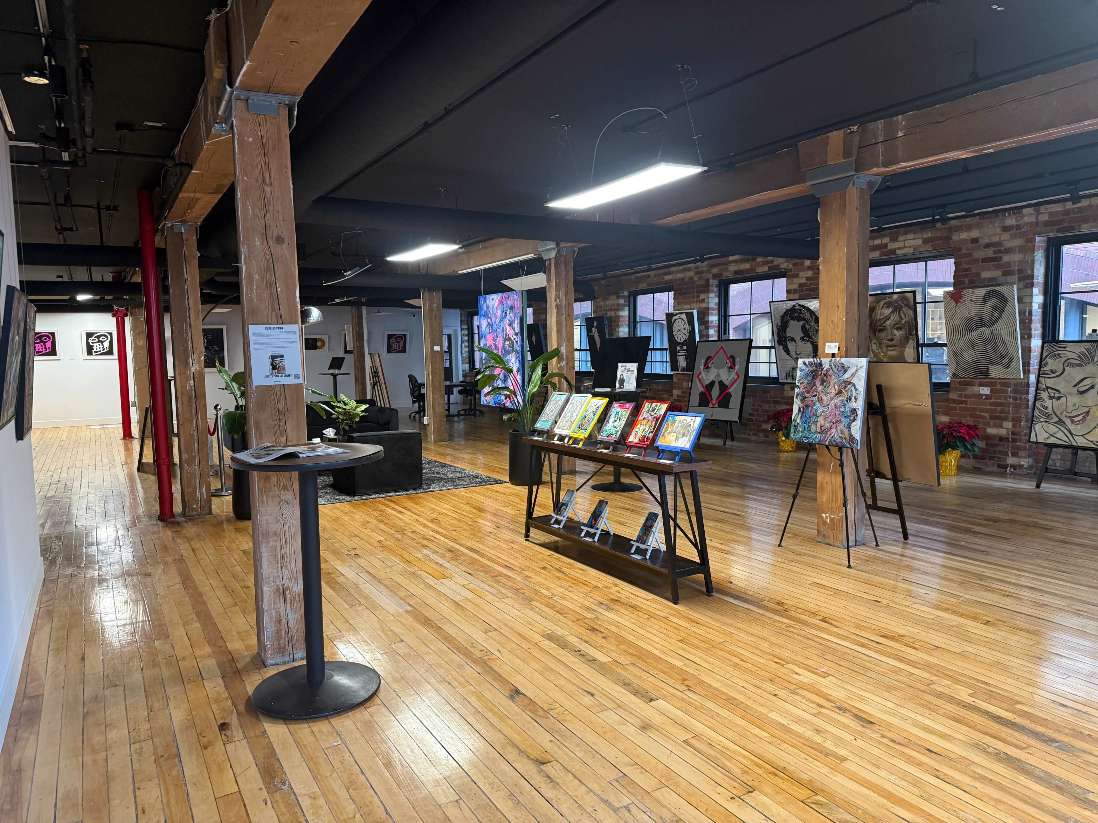 The image shows an art gallery with wooden floors, exposed brick walls, and various artworks displayed on stands and walls.