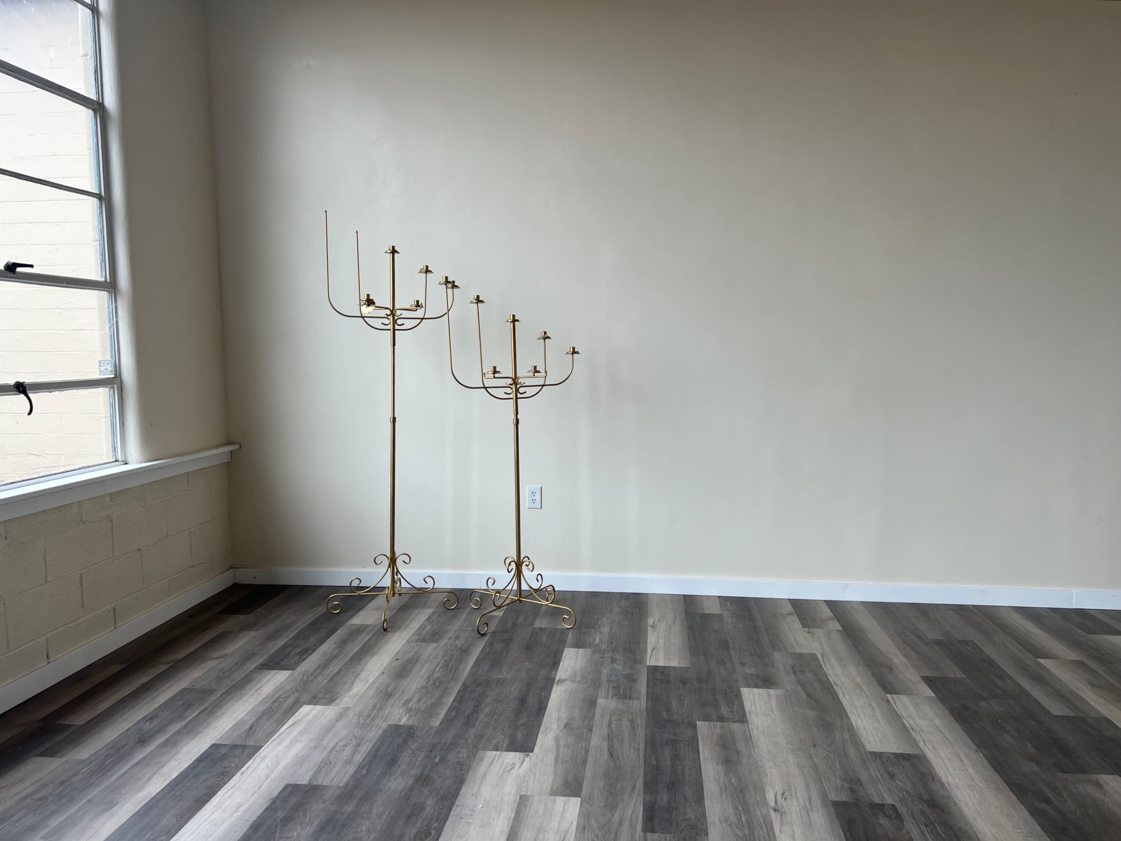 Two golden candle holders stand in an empty room with light-colored walls and wood-like flooring.
