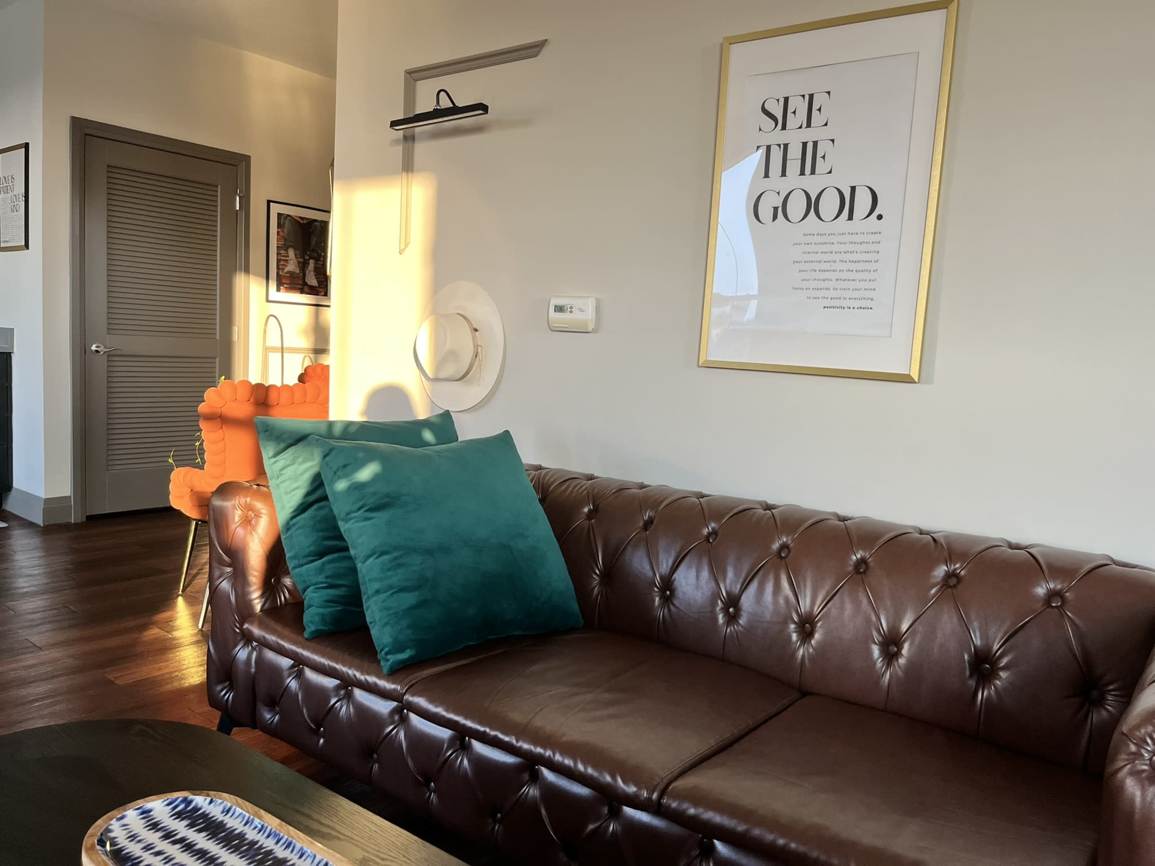 A brown leather couch with green cushions is positioned in a well-lit living room featuring a framed poster on the wall and an orange chair in the background.
