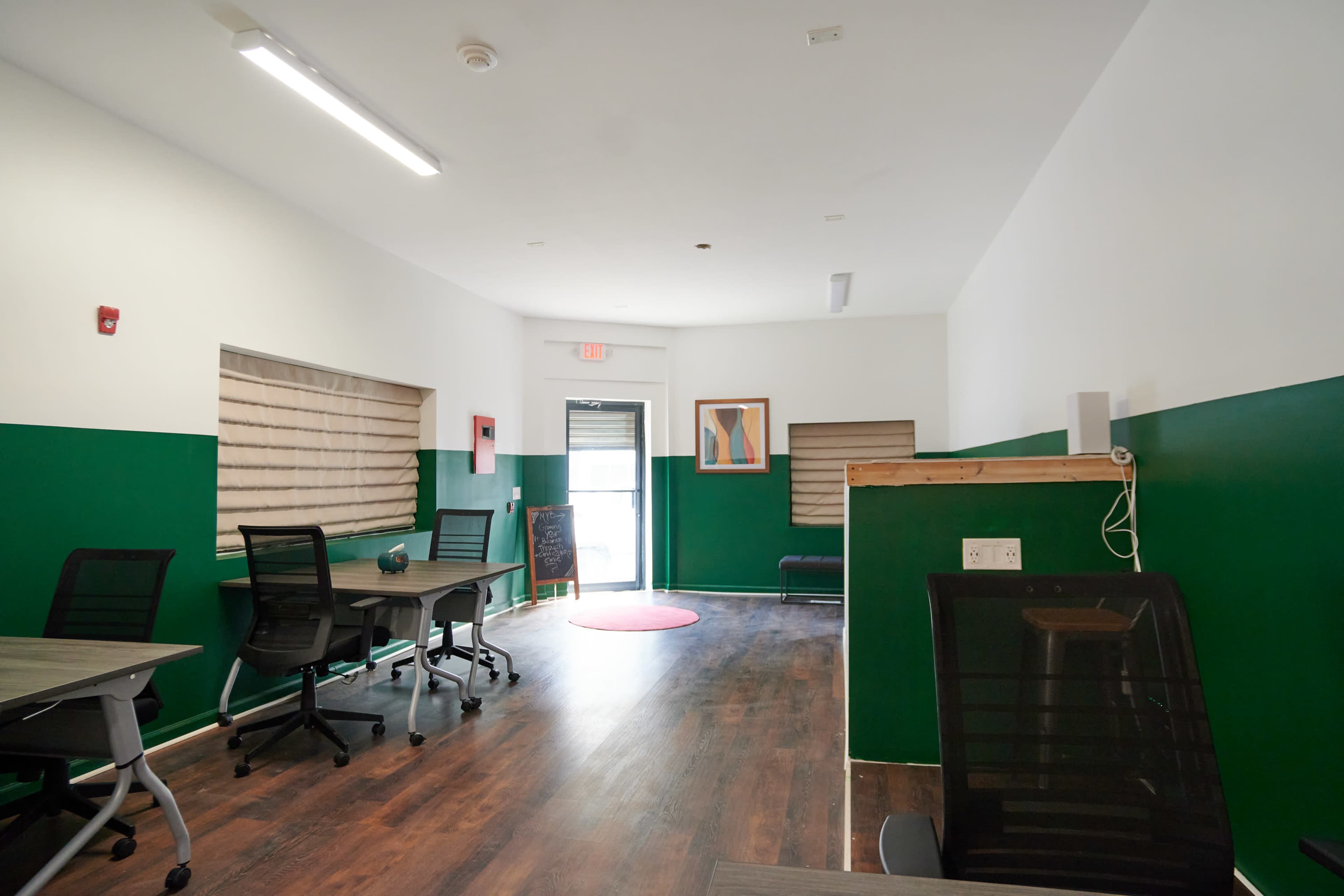 A modern office space with green and white walls, featuring several desks and a door leading outside.