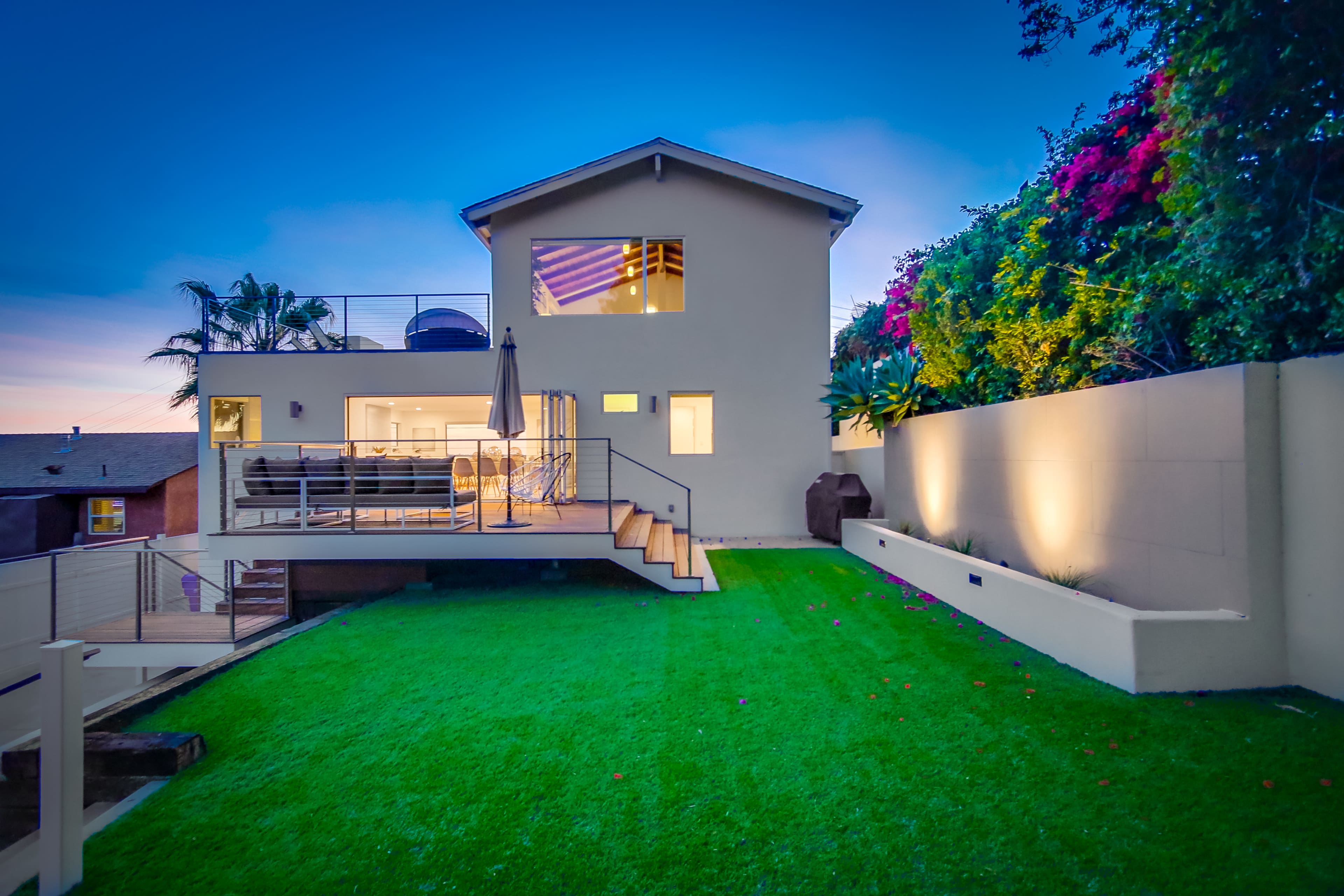 A modern two-story house with a patio and green lawn is set against a backdrop of colorful foliage during twilight.