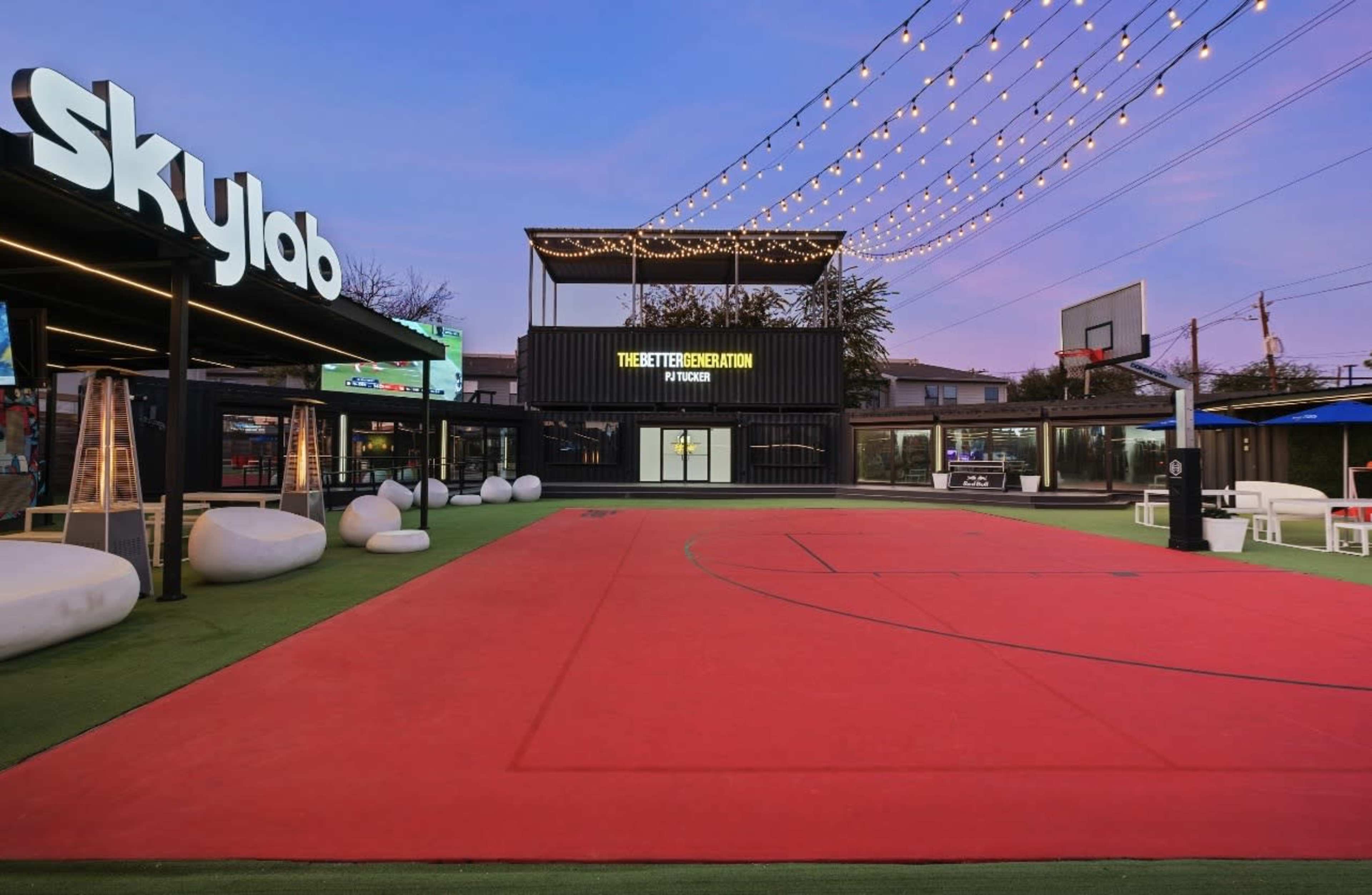 The image shows a sports facility featuring a red basketball court, a black building with "Skylab" signage, and string lights overhead in an outdoor setting.